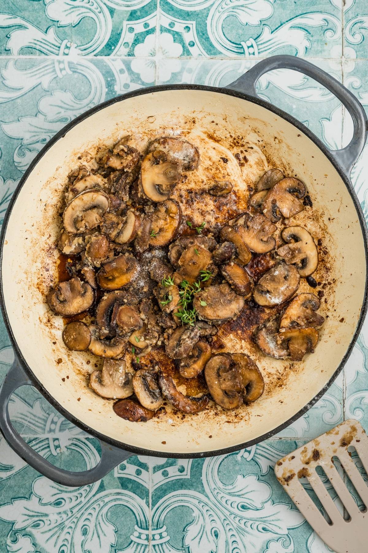 Sliced mushrooms cooking in a large white skillet with herbs, on a patterned blue tile background.