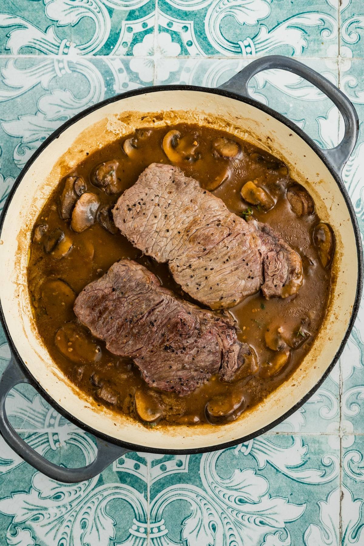 Two cooked steaks in a brown mushroom sauce in a round pot on a blue and white patterned surface.