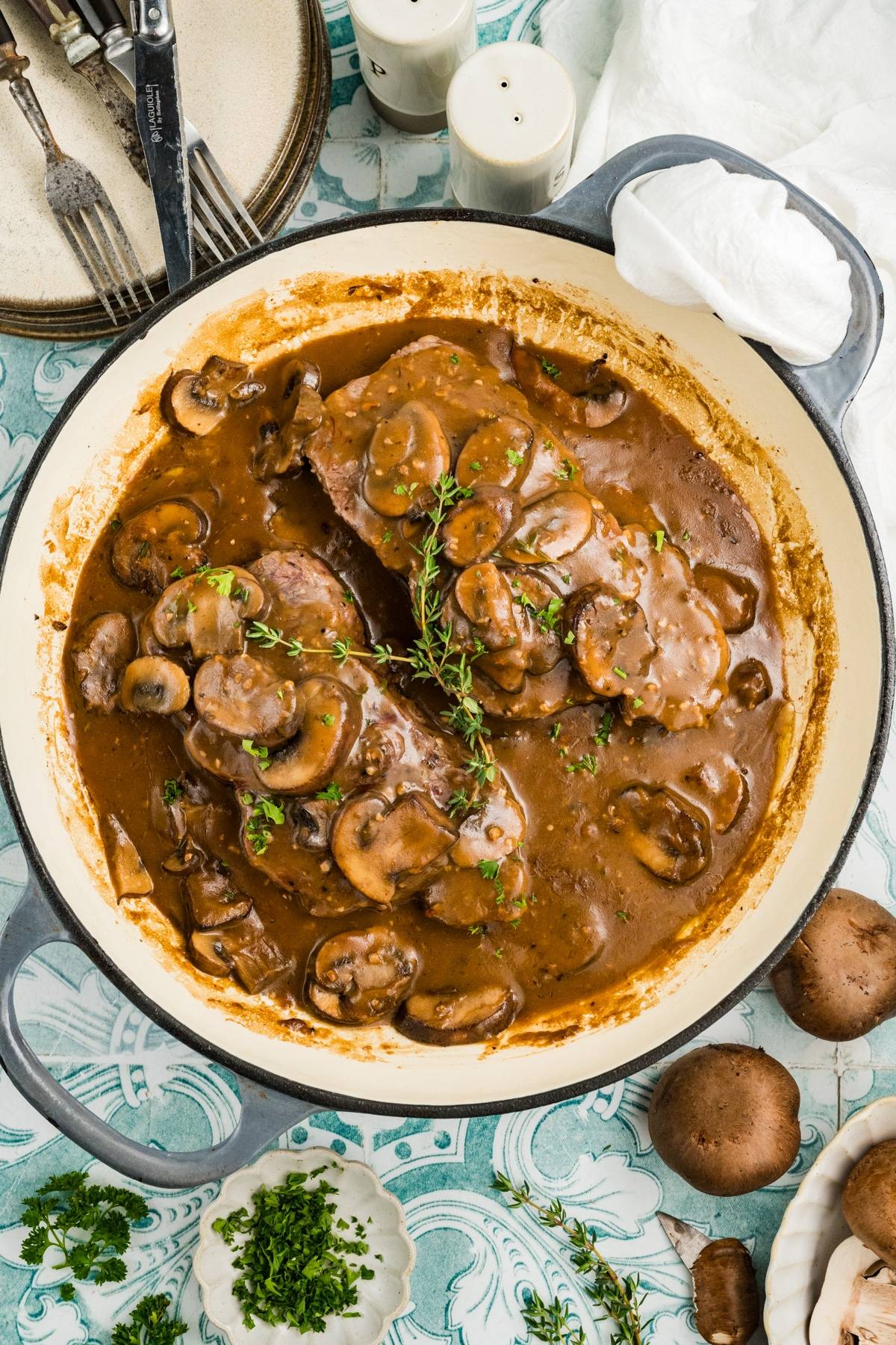 Pot of marsala steak in mushroom gravy, garnished with herbs, surrounded by mushrooms and fresh parsley.