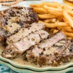 Sliced steak au poivre with creamy peppercorn sauce and a side of golden French fries on a green plate.