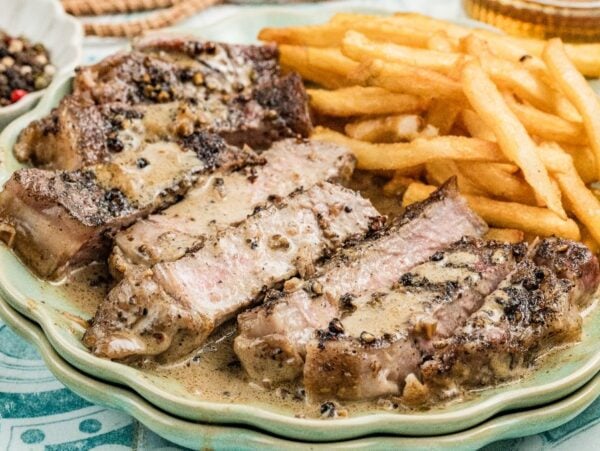 Sliced steak au poivre with creamy peppercorn sauce and a side of golden French fries on a green plate.