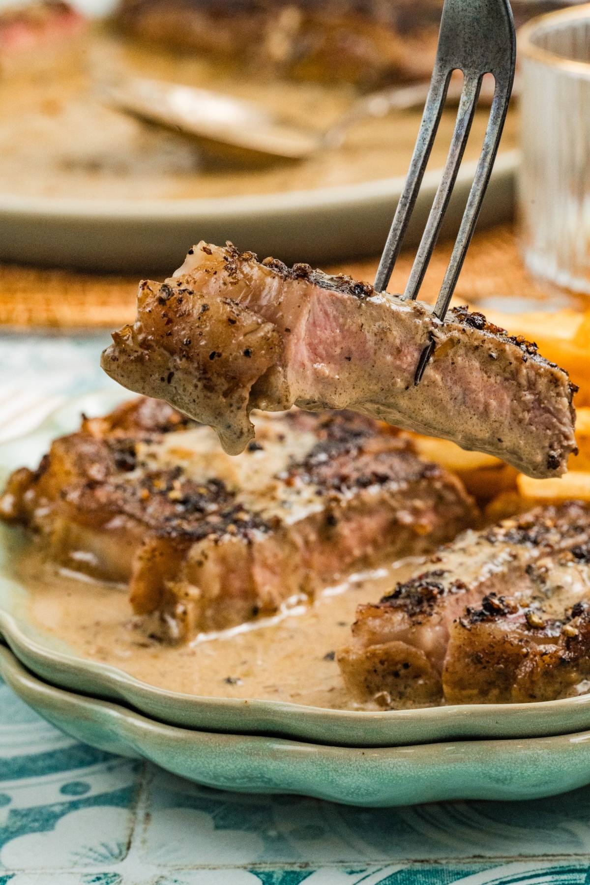 A fork holds a juicy, peppered steak slice above a plate with steak in creamy sauce.