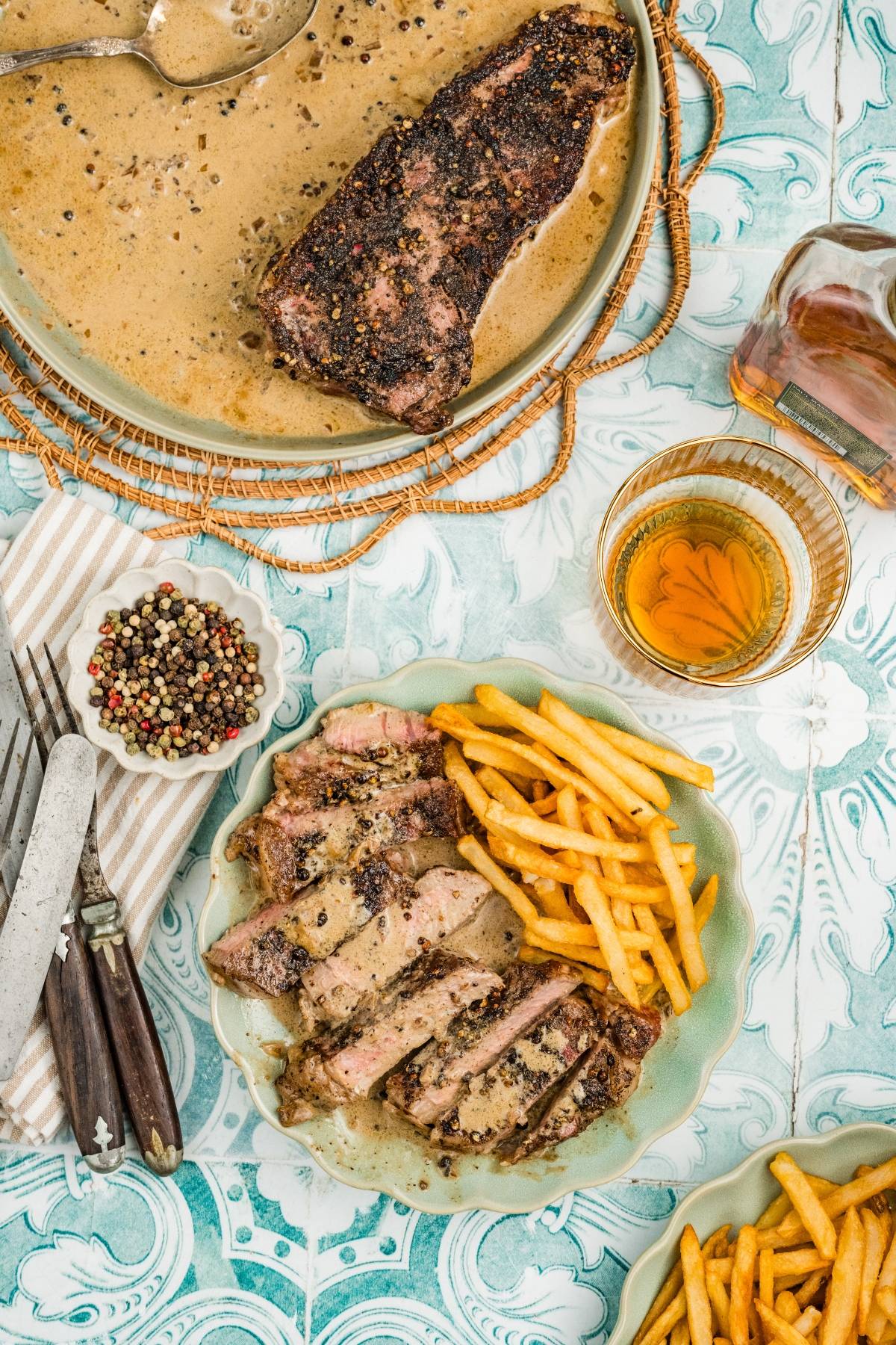 Sliced steak with peppercorn sauce, French fries, and a drink on a patterned table with cutlery and peppercorns.