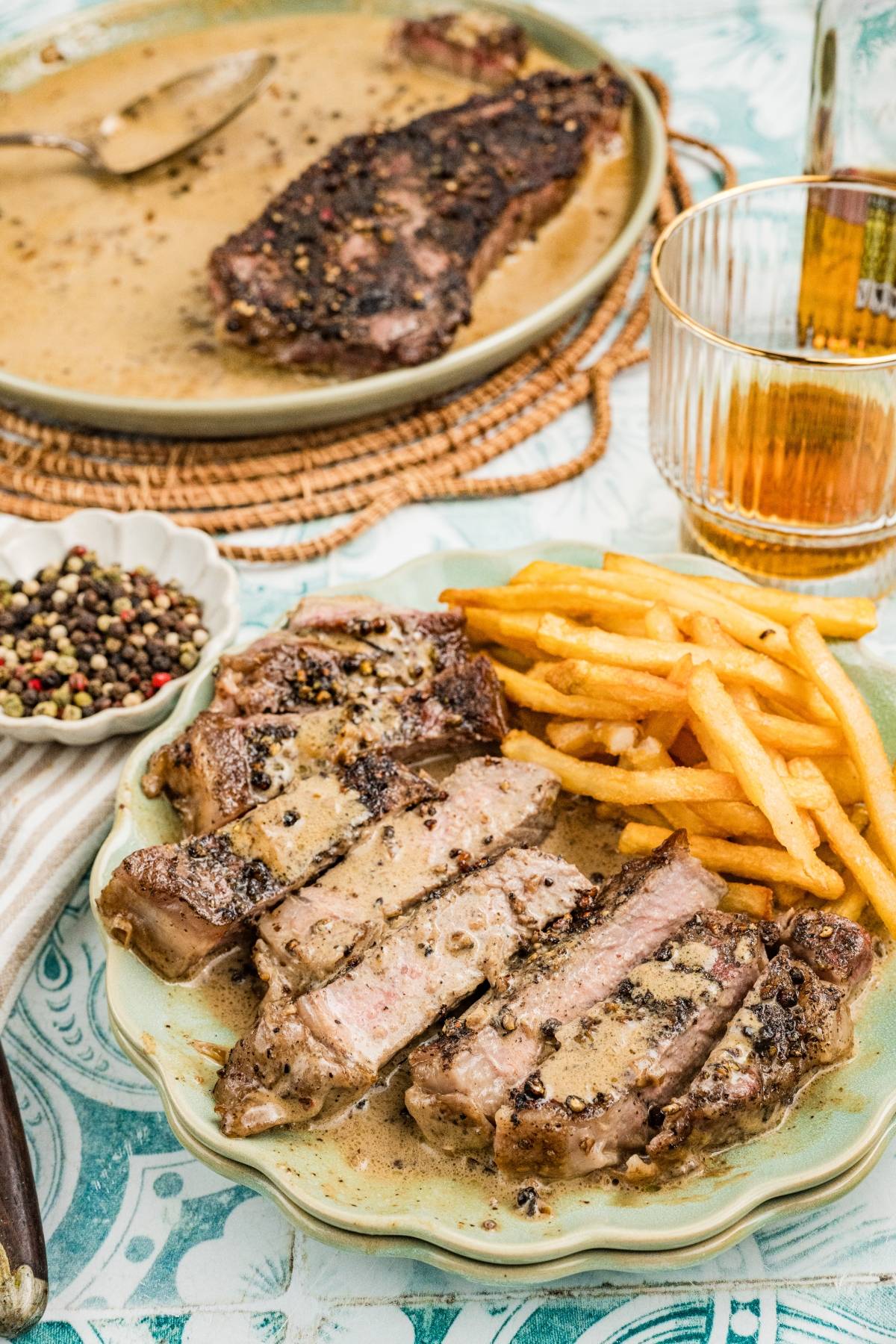 Sliced steak with creamy peppercorn sauce and fries on a plate, with a drink and whole steak in the background.
