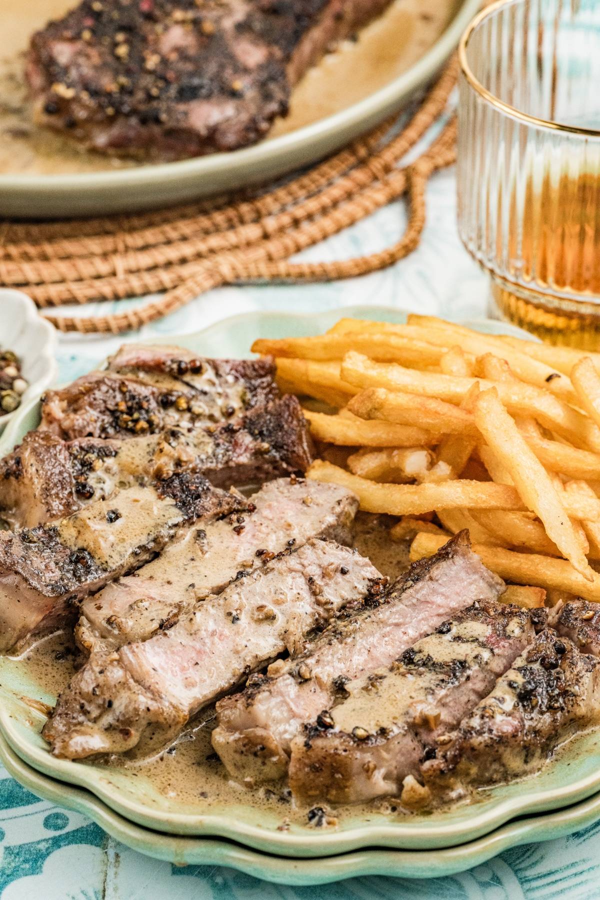 Sliced steak with creamy pepper sauce and French fries on a plate, with a drink in the background.