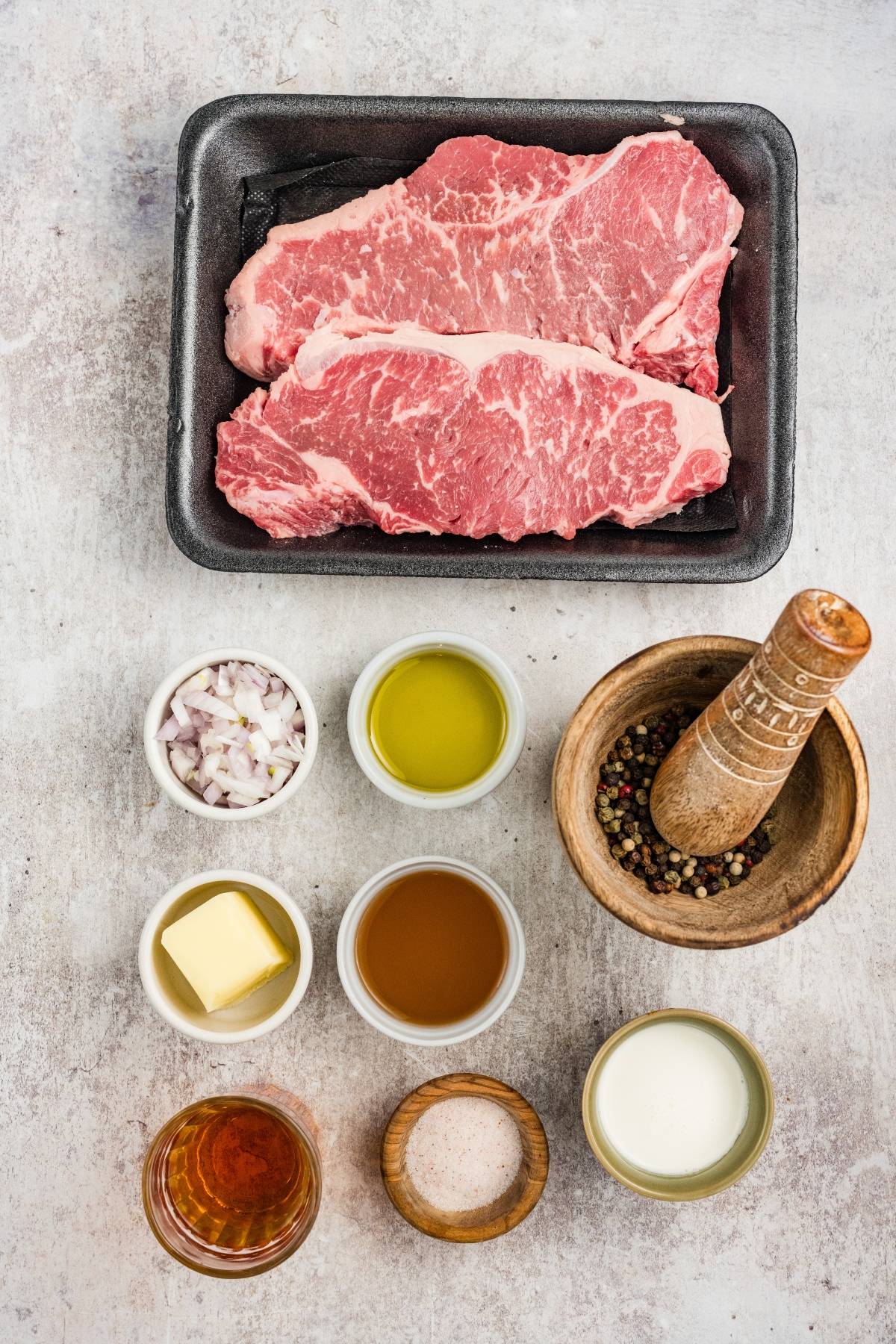 Two raw steaks in a tray with small bowls of oil, shallots, butter, vinegar, cream, salt, and peppercorns.
