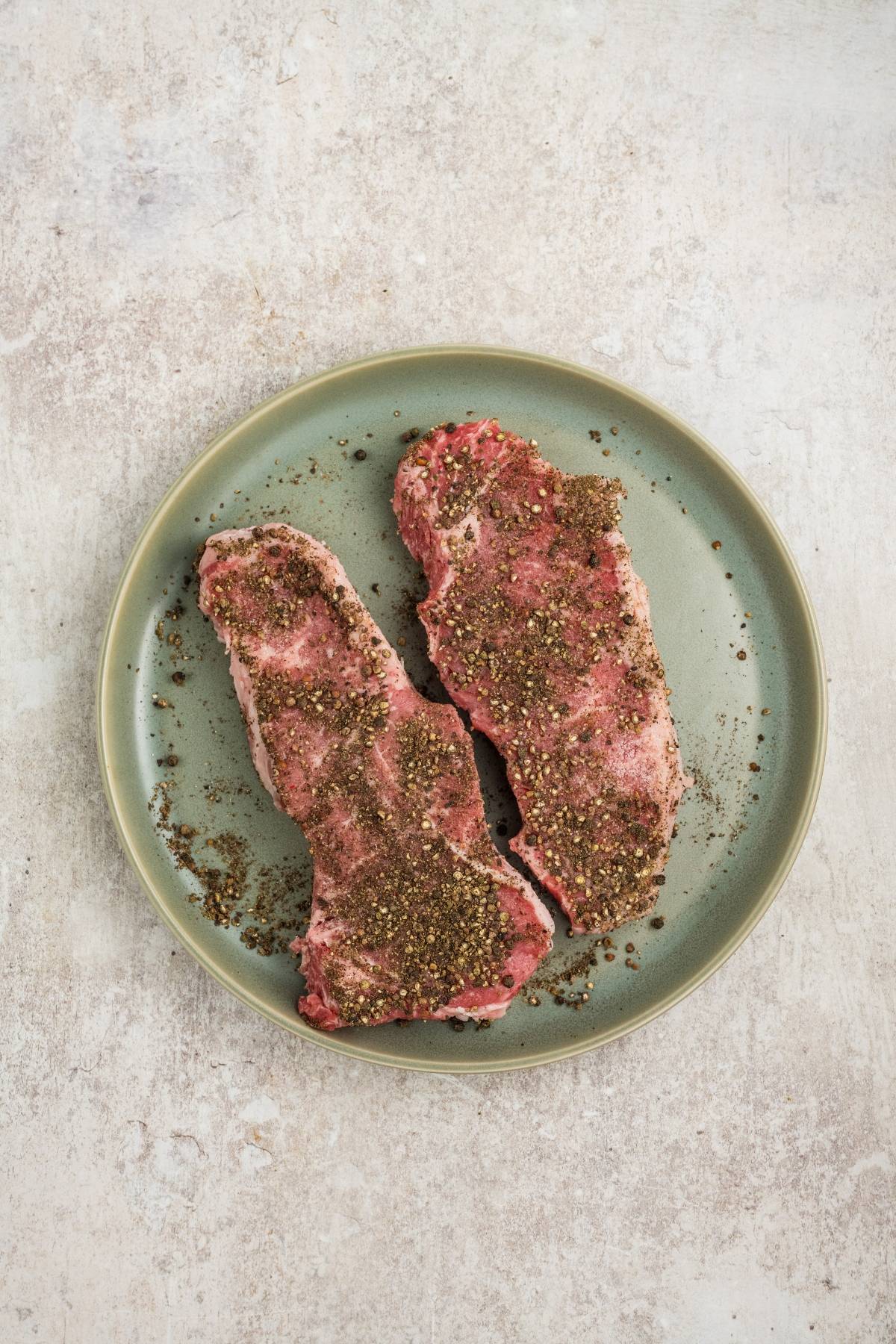 Two raw, seasoned steaks on a green plate atop a light, textured surface.