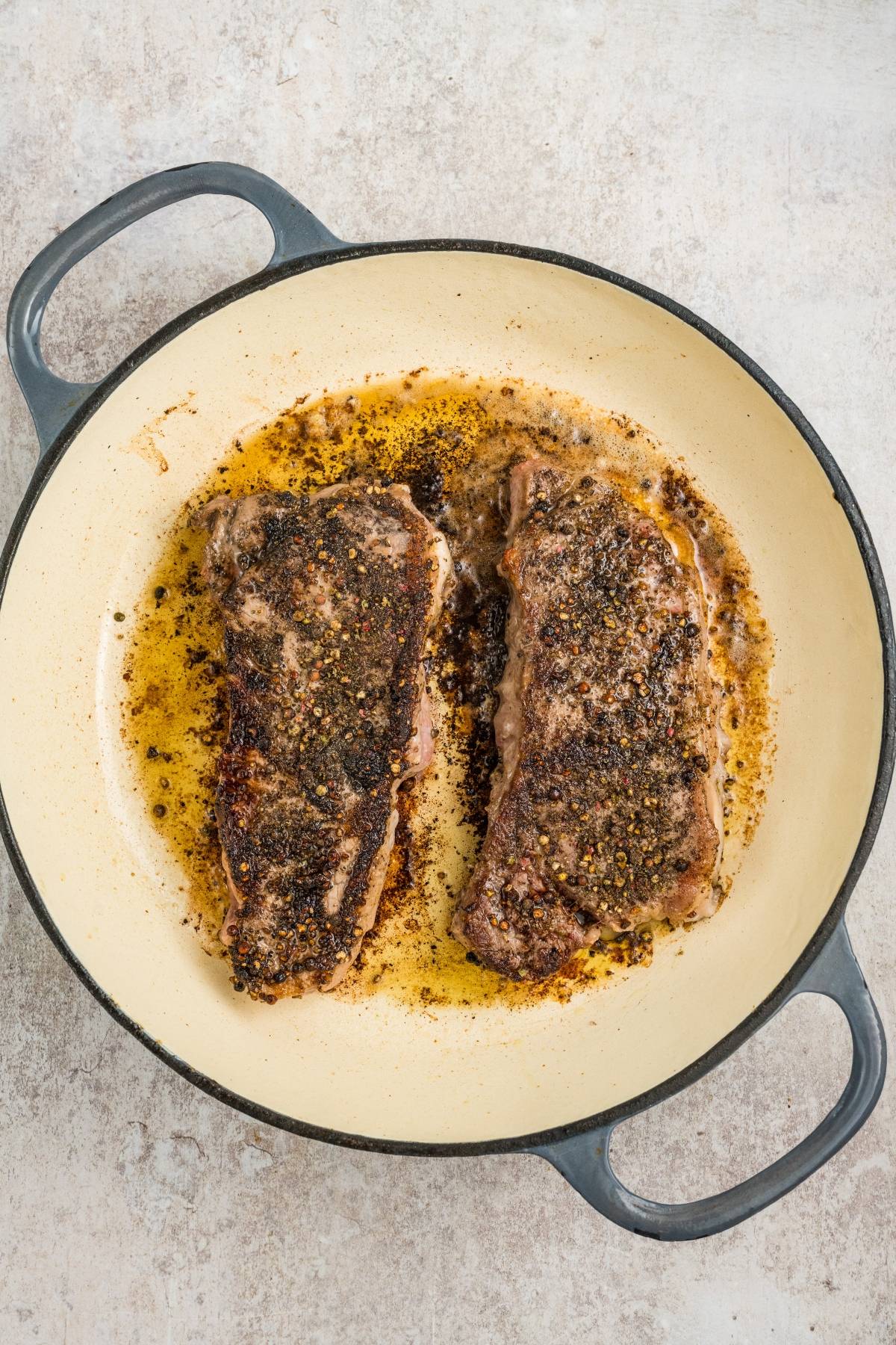Two cooked steaks with seasoning in a round cream-colored pan with handles, sitting in melted butter and herbs.