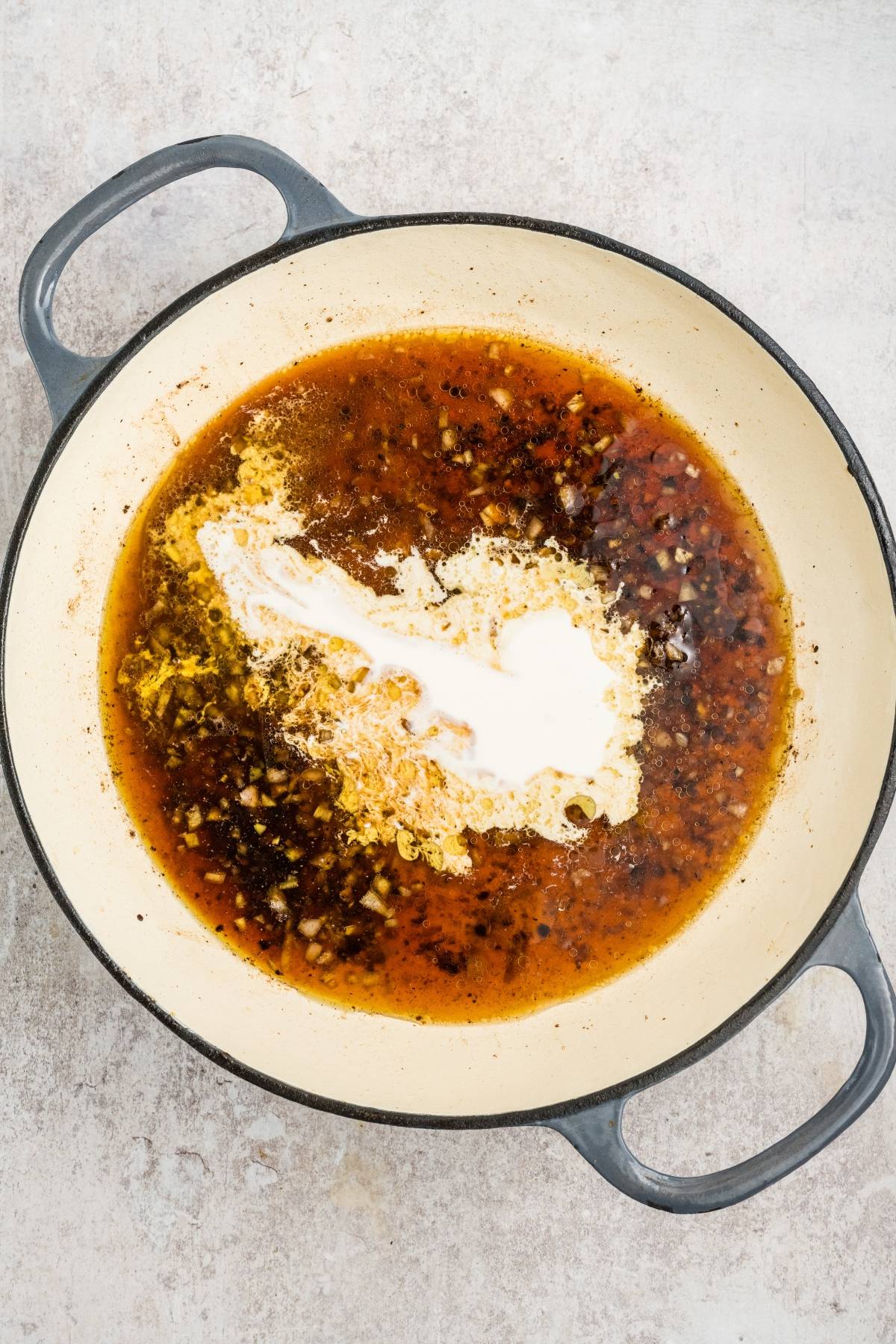 Large pot with simmering brown sauce, chopped garlic, and a swirl of cream on a light-colored surface.