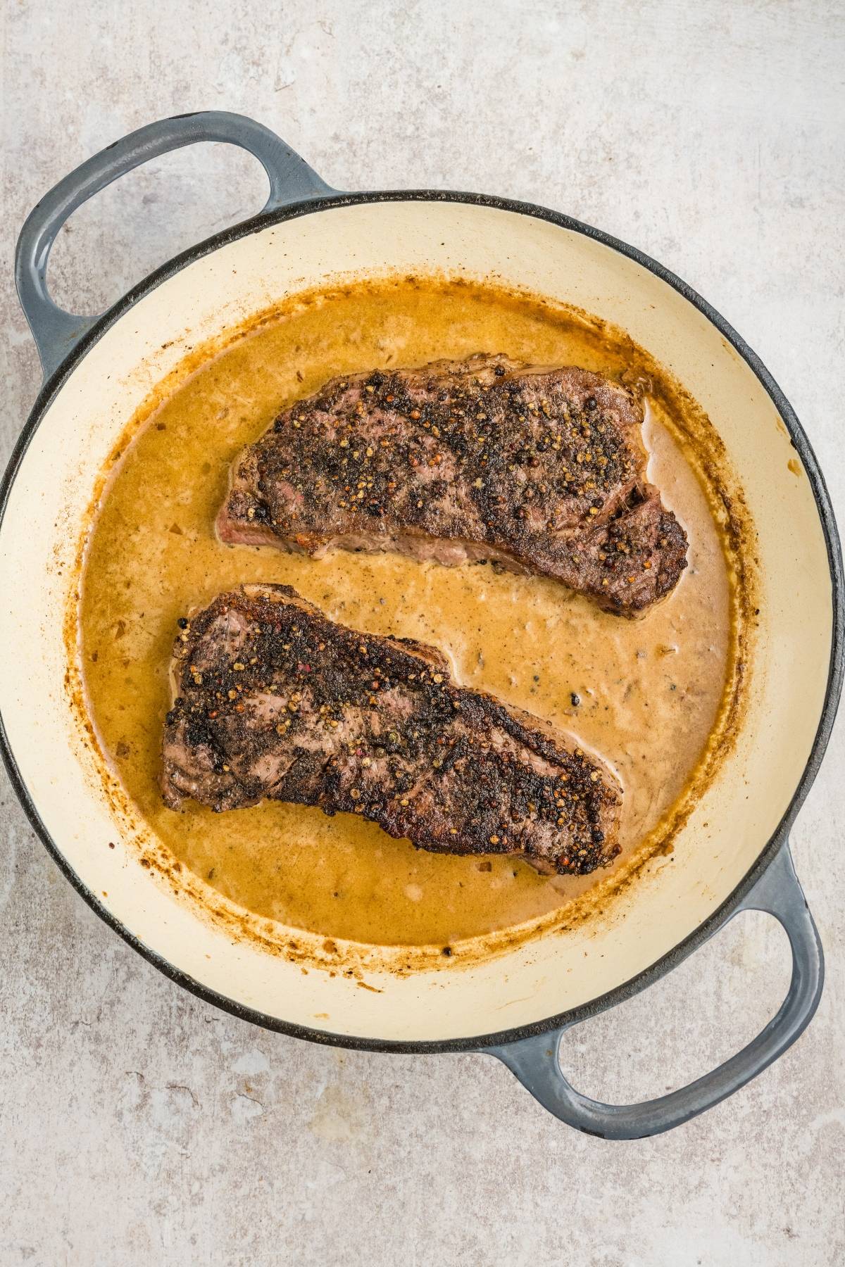Two cooked steaks in a creamy brown sauce inside a round enameled cast iron pot on a light surface.