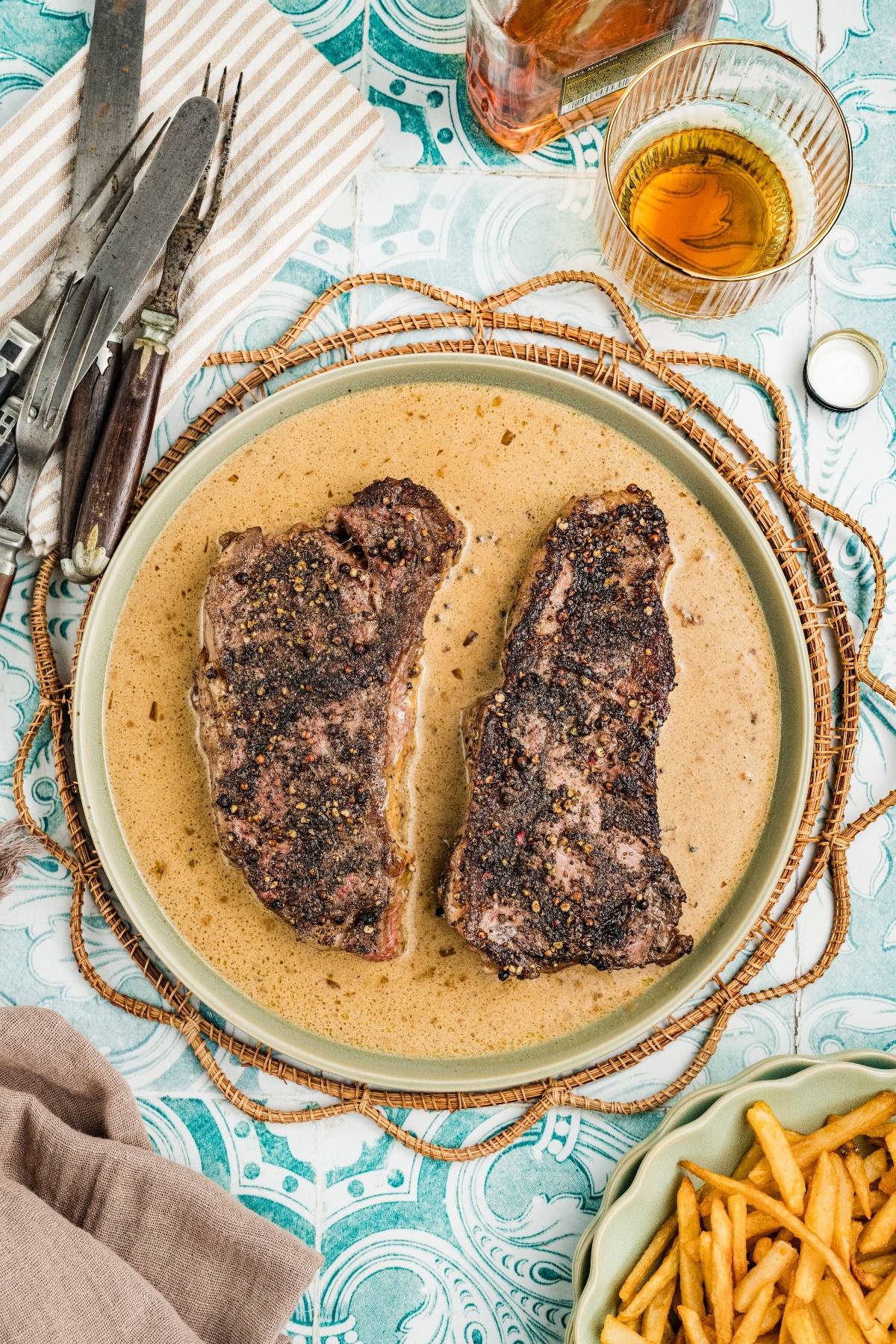 Two seasoned steaks on a plate with creamy sauce, next to fries, cutlery, and a glass of amber drink.