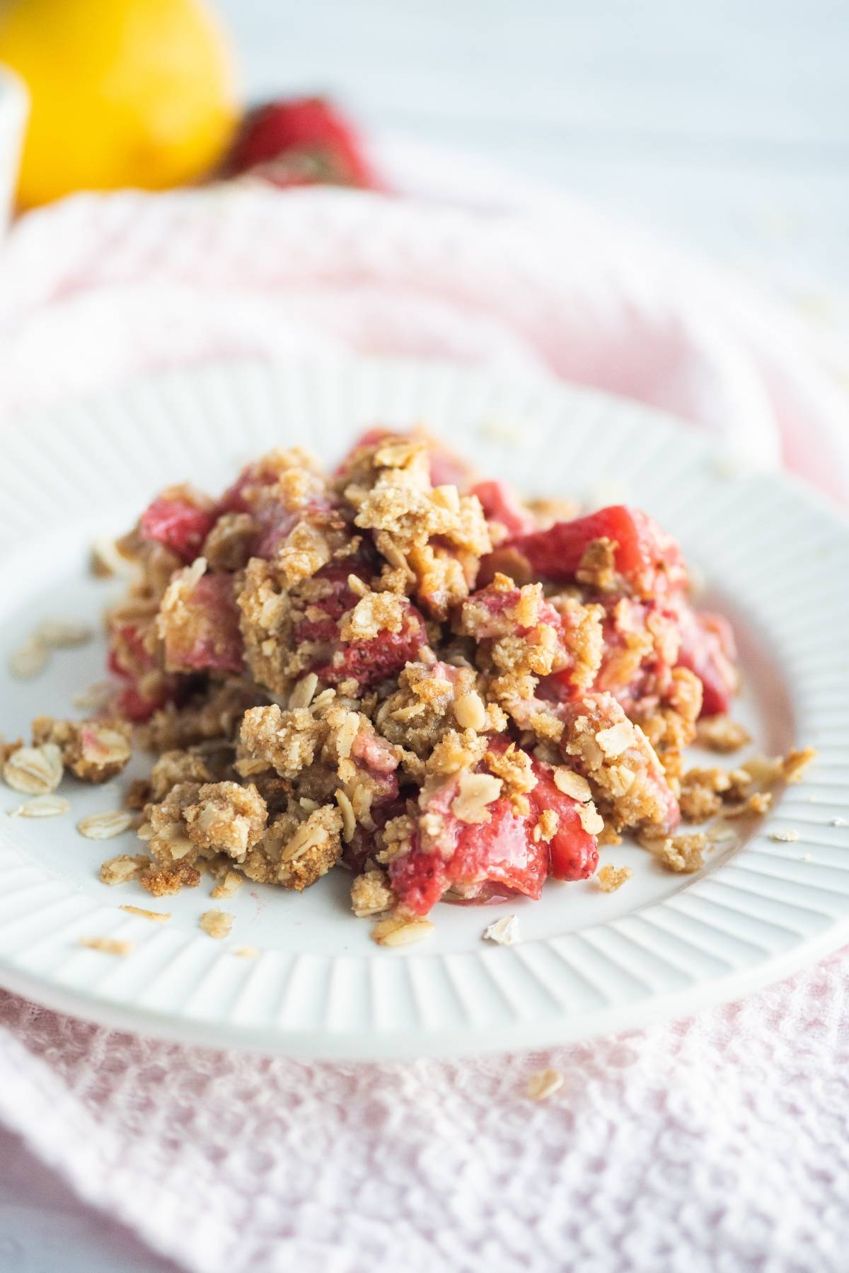 A serving of strawberry crumble or crisp with oat topping sits on a white plate, placed on a pink cloth—an inviting dessert made with fresh berries.