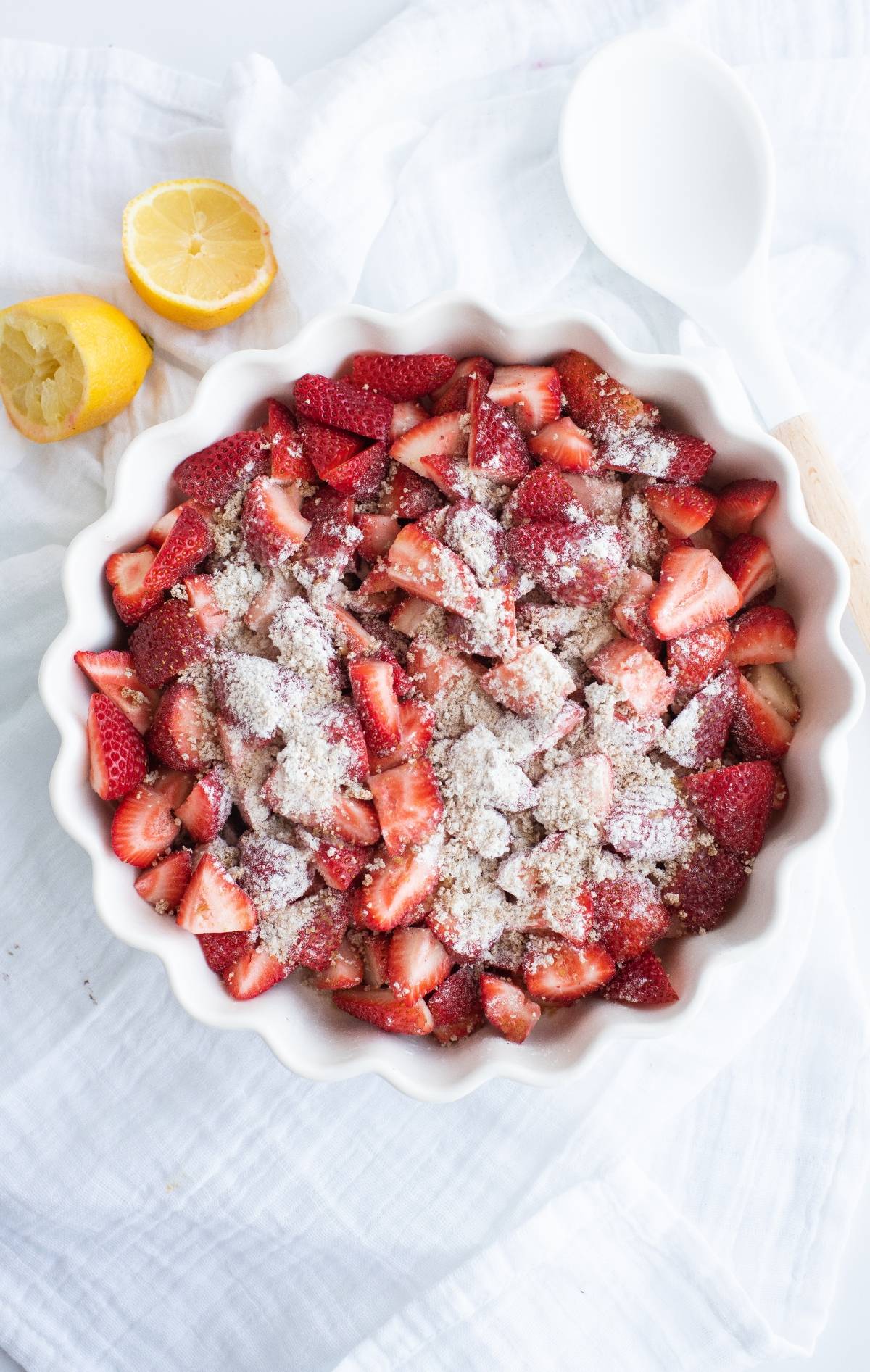Sliced strawberries in a white dish topped with flour, beside a halved lemon and white spoon—perfect for making a Strawberry Crisp, the best gluten free crisp recipe for a delicious baked dessert with fruit.
