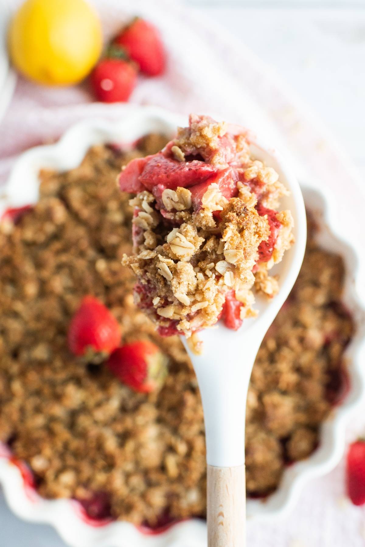 A white spoon holds a portion of strawberry crumble or crisp above a pie dish, with fresh strawberries nearby—a delightful dessert made with fresh berries.