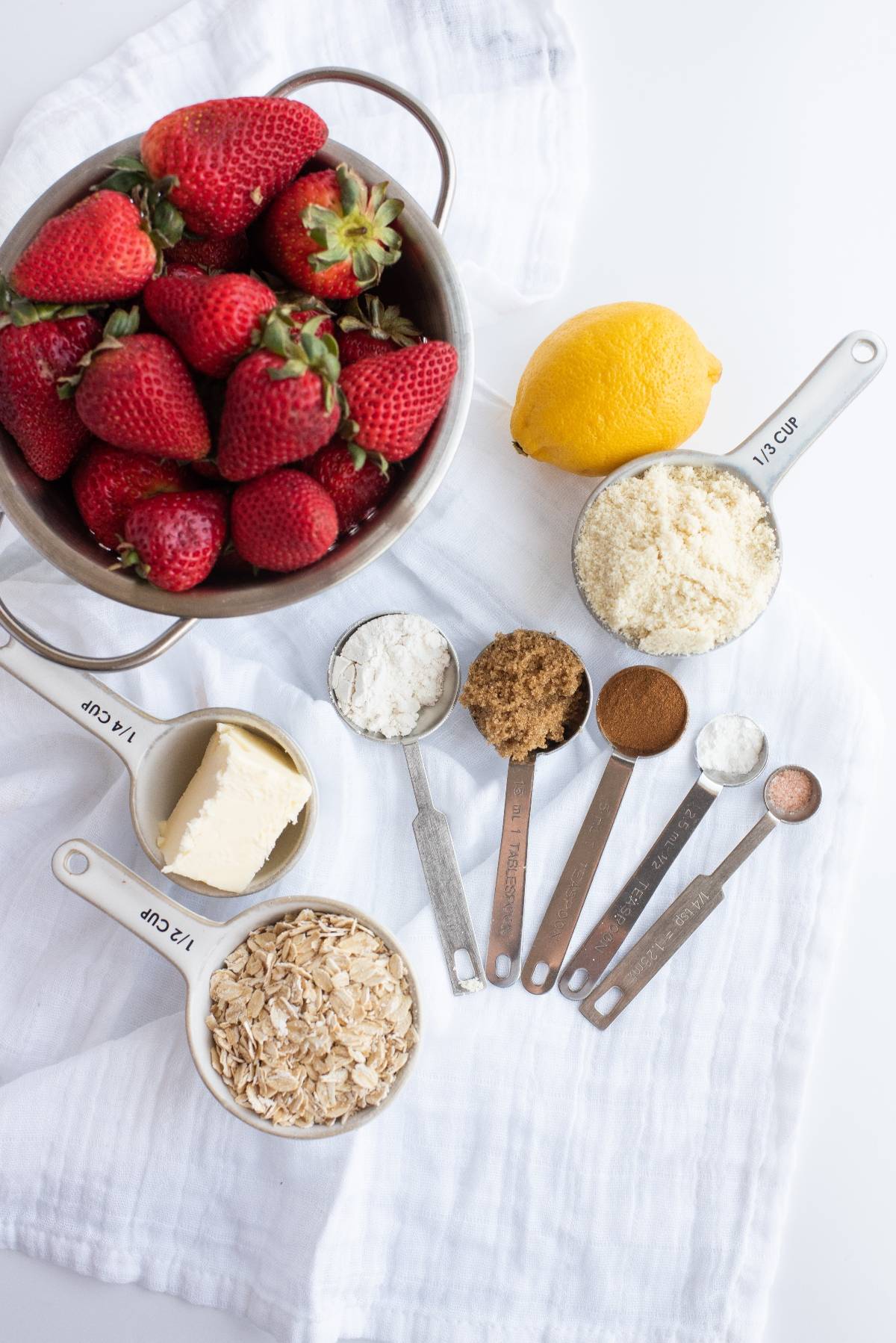 Fresh strawberries, a lemon, butter, oats, flour, almond flour, brown sugar, and spices are ready to become a homemade strawberry crumble or crisp on a white cloth.