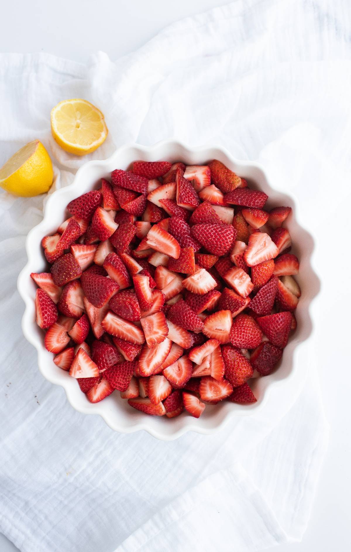 Bowl filled with sliced strawberries, perfect for a refreshing strawberry crisp, sits next to a halved lemon on a white cloth.