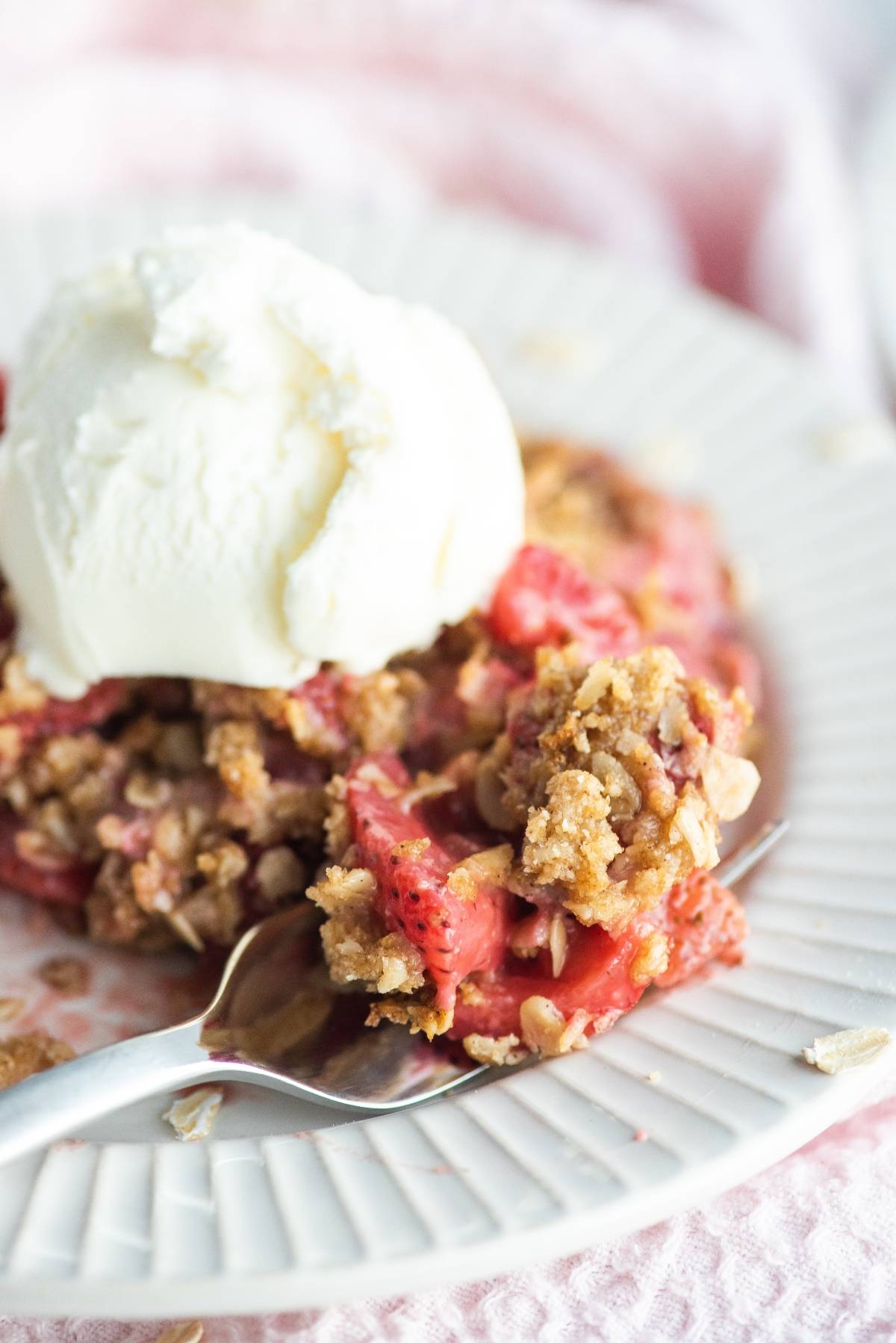A close-up of strawberry crisp, a classic baked dessert with fruit, served with a scoop of vanilla ice cream on a white plate and a spoon.