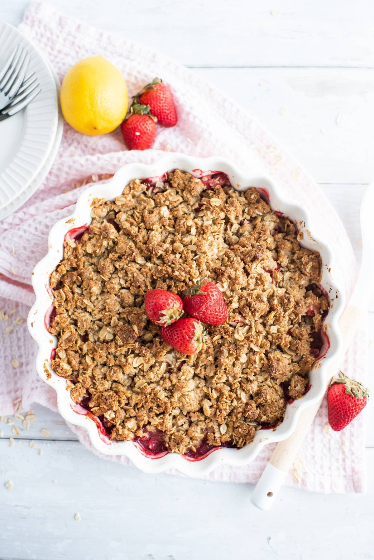 A baked strawberry crisp in a white pie dish, topped with fresh strawberries on a white table—this is the best gluten free crisp recipe for a delicious dessert made with fresh berries.
