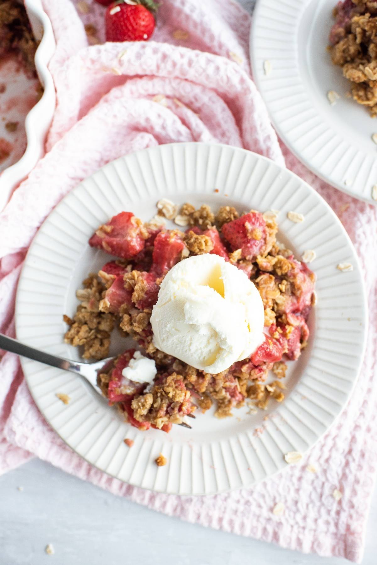 A plate of strawberry crumble or crisp topped with a scoop of vanilla ice cream sits on a white plate with a fork, making for the perfect baked dessert with fruit.
