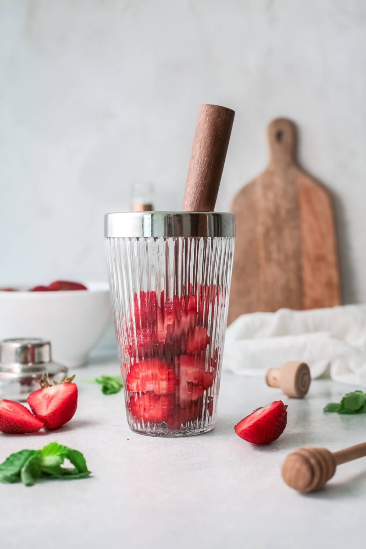 A glass for a refreshing strawberry tequila cocktail with sliced strawberries and a muddler, surrounded by mint leaves and honey dippers on the countertop.