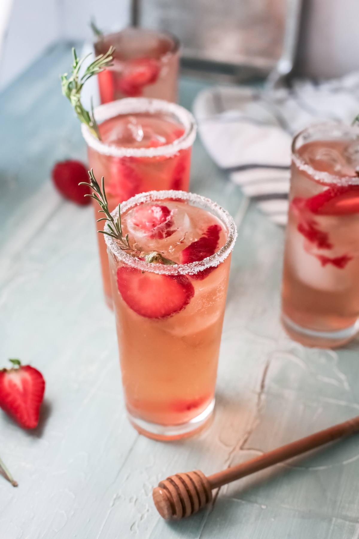 Tall glasses of strawberry tequila cocktail with ice, sugar rims, and rosemary sprigs sit on a light blue table.