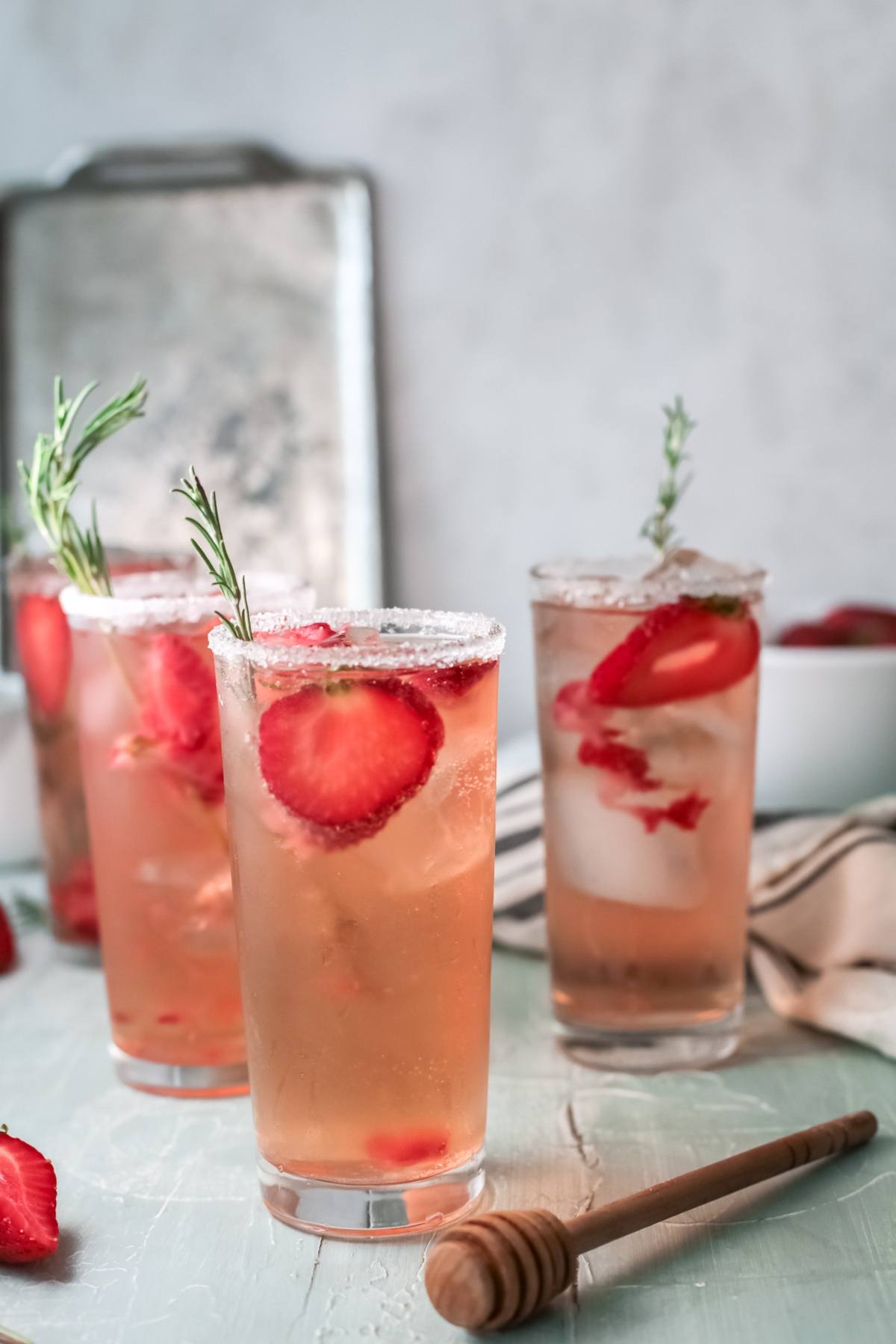 Three tall glasses of a cocktail with strawberries and ice, garnished with rosemary, sit on a light table with a honey dipper nearby.