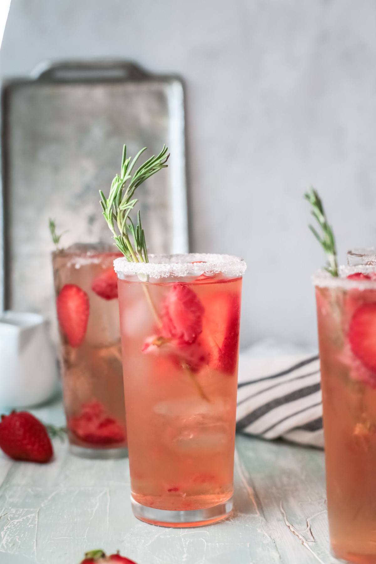 A tall glass of pink strawberry paloma with ice and rosemary garnish, rimmed with sugar—a refreshing cocktail with strawberries perfect for summer.