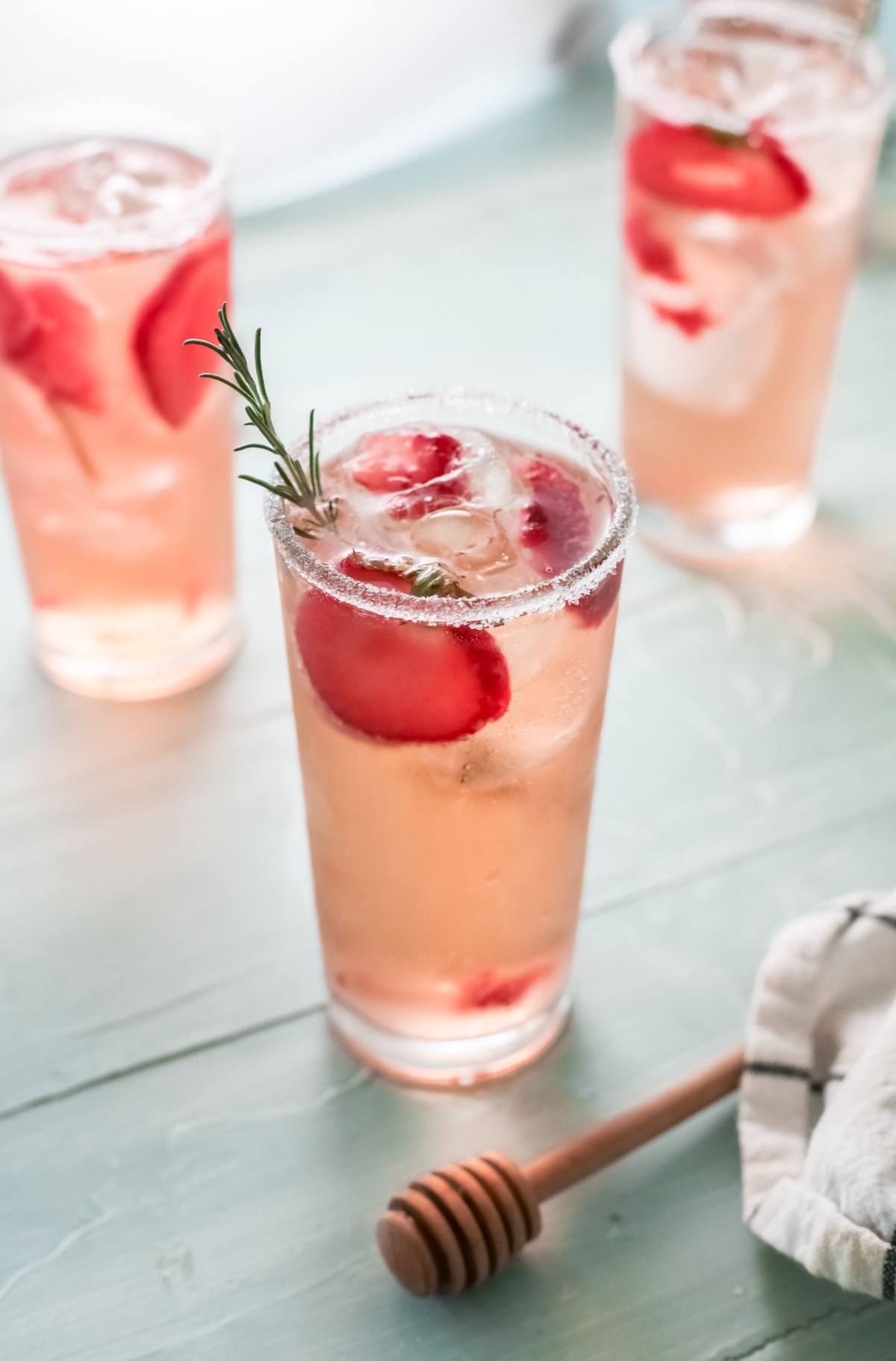 Three tall glasses of pink Strawberry Paloma with ice, strawberry slices, and rosemary sprigs sit on a light blue surface—an inviting cocktail with strawberries perfect for any occasion.