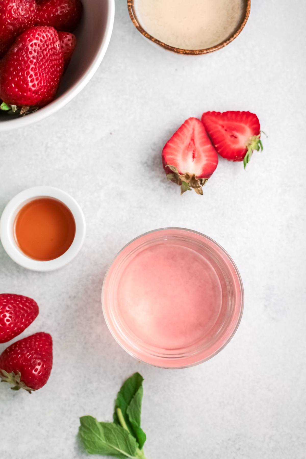 Glass of pink strawberry smoothie surrounded by strawberries, mint, and small bowls of ingredients on a light surface, perfect as inspiration for an Easy paloma recipe or a refreshing strawberry tequila cocktail.