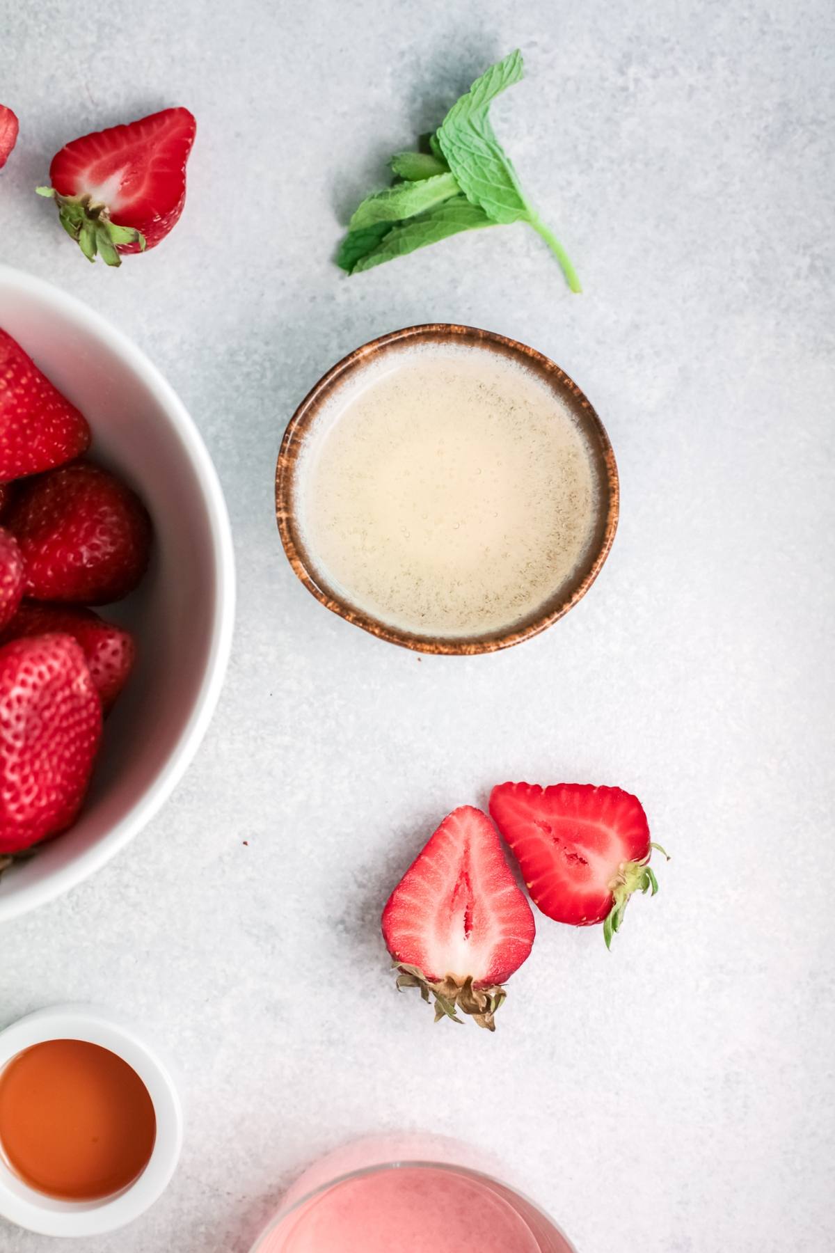 Bowl of strawberries, a small cup of sauce, mint sprig, and strawberry halves on a light surface—perfect garnishes for your next strawberry tequila cocktail.