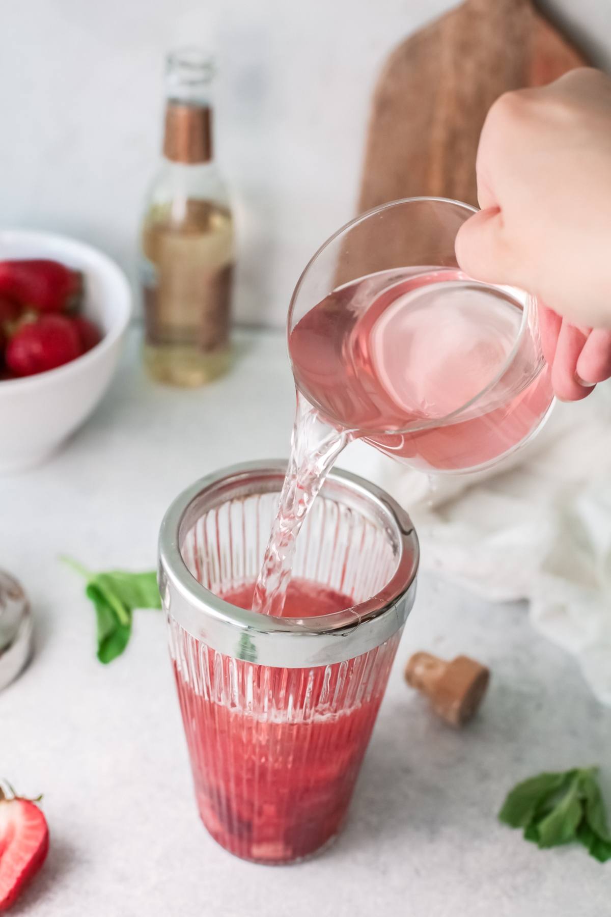 A hand pours clear liquid into a glass with red juice to craft a refreshing strawberry tequila cocktail; strawberries and a bottle are nearby.