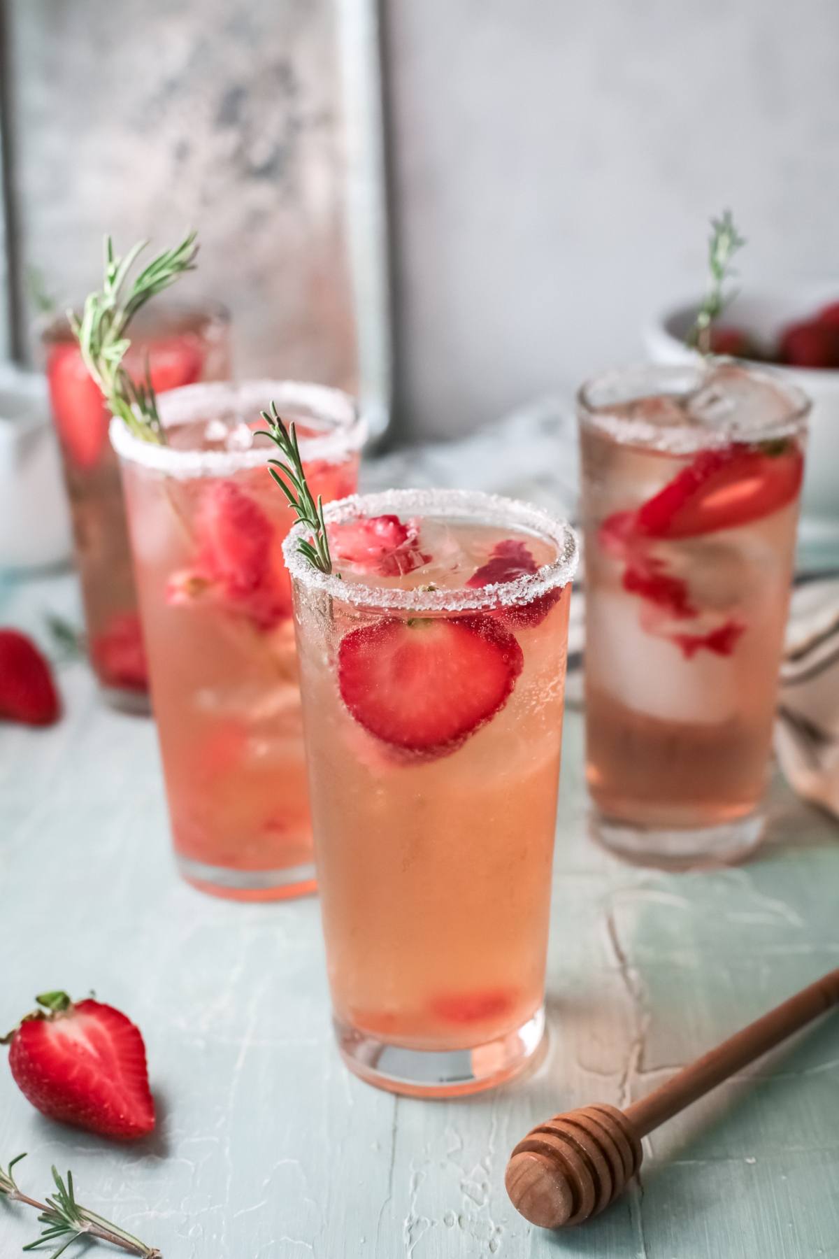 Three tall glasses of a refreshing strawberry tequila cocktail with ice, garnished with rosemary and sugar rims, on a light table.
