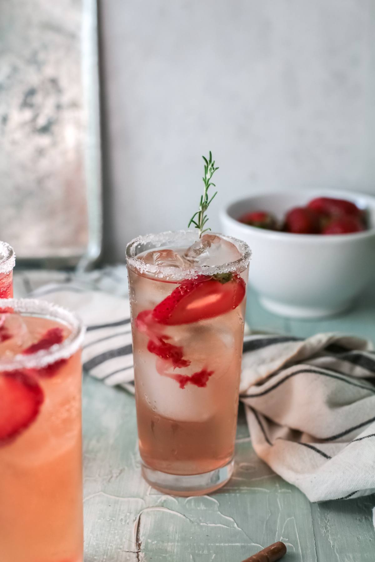 A glass of iced strawberry tequila cocktail garnished with a sprig of thyme, next to a bowl of strawberries.