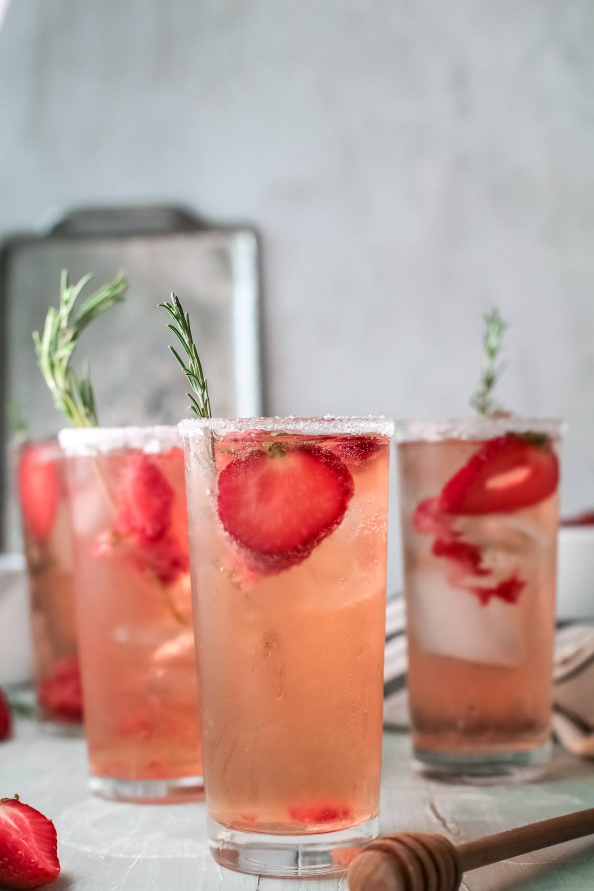 Iced strawberry paloma drinks with rosemary sprigs in tall glasses, garnished with sugar rims, on a light surface—a refreshing and easy paloma recipe perfect for summer.