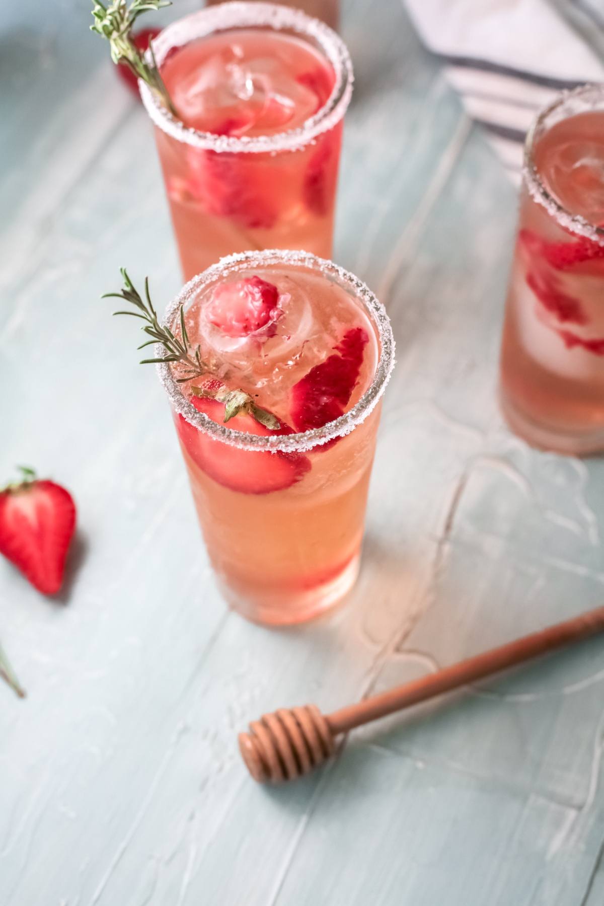 Iced strawberry drinks in tall glasses, garnished with rosemary and strawberries, on a light blue surface—an easy paloma recipe perfect for summertime sipping.