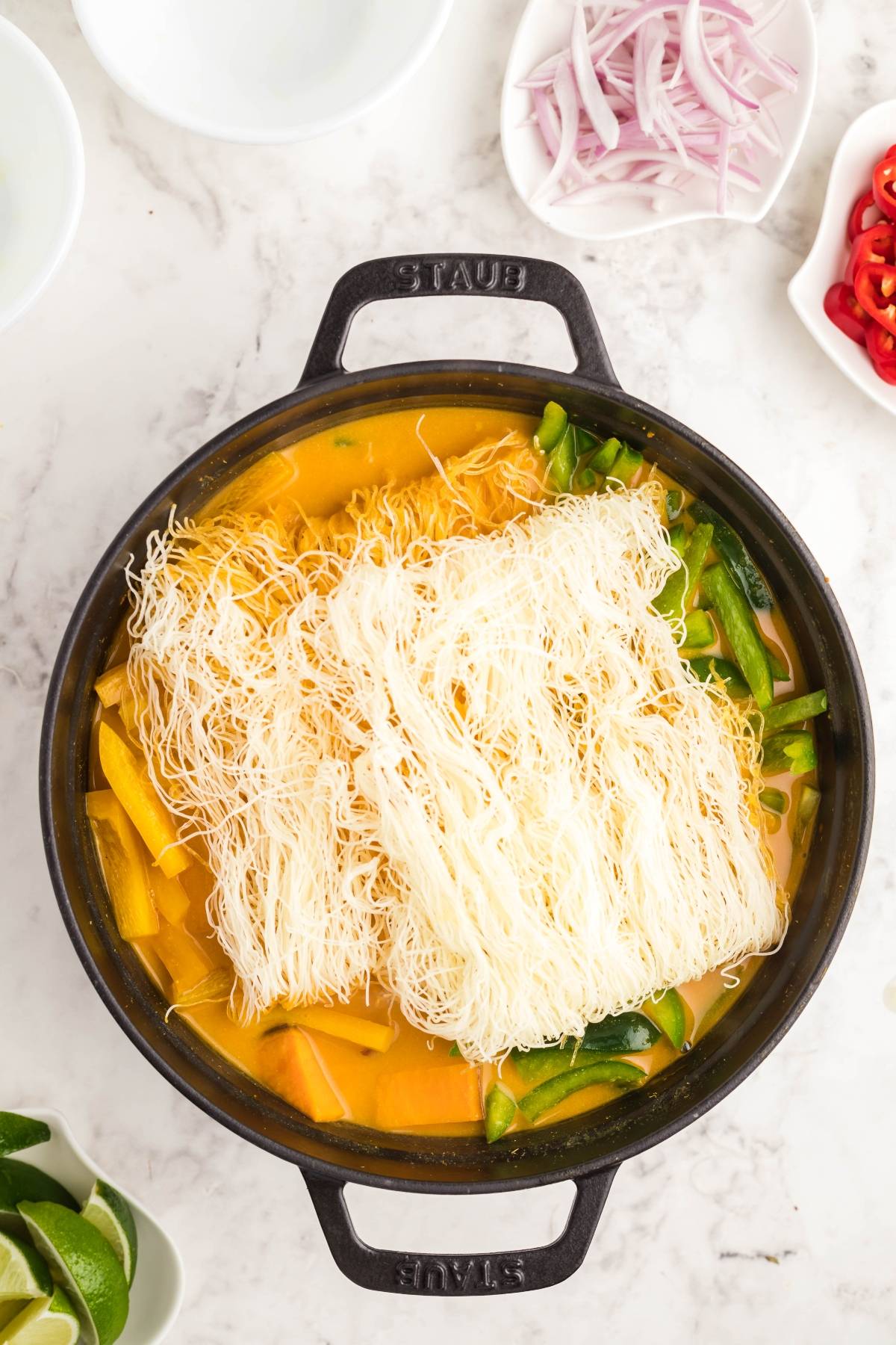 A pot of hearty Thai recipe with chicken, dry rice noodles, vegetables, and broth, surrounded by sliced onions, chilies, and lime wedges.