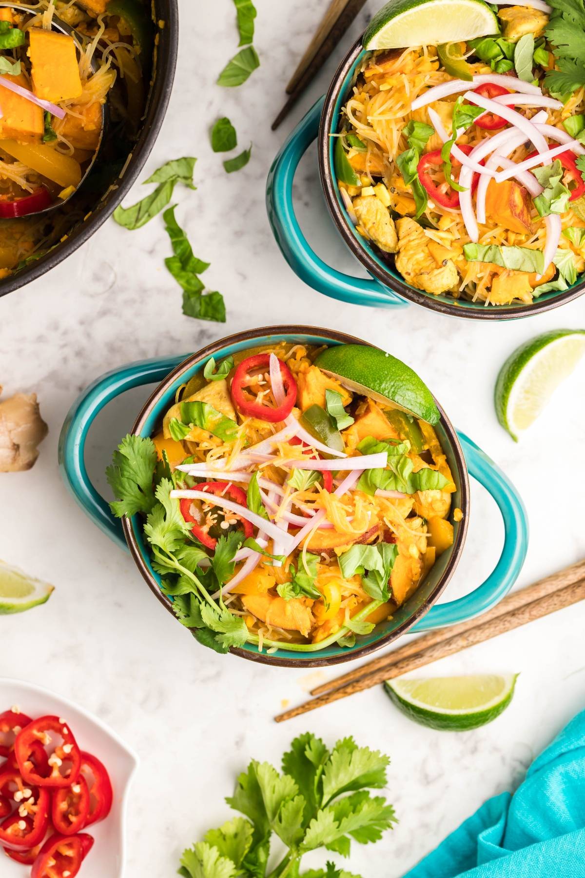 Two bowls of vibrant soup with red curry, topped with cilantro, red onion, chilies, and lime slices on a white surface.