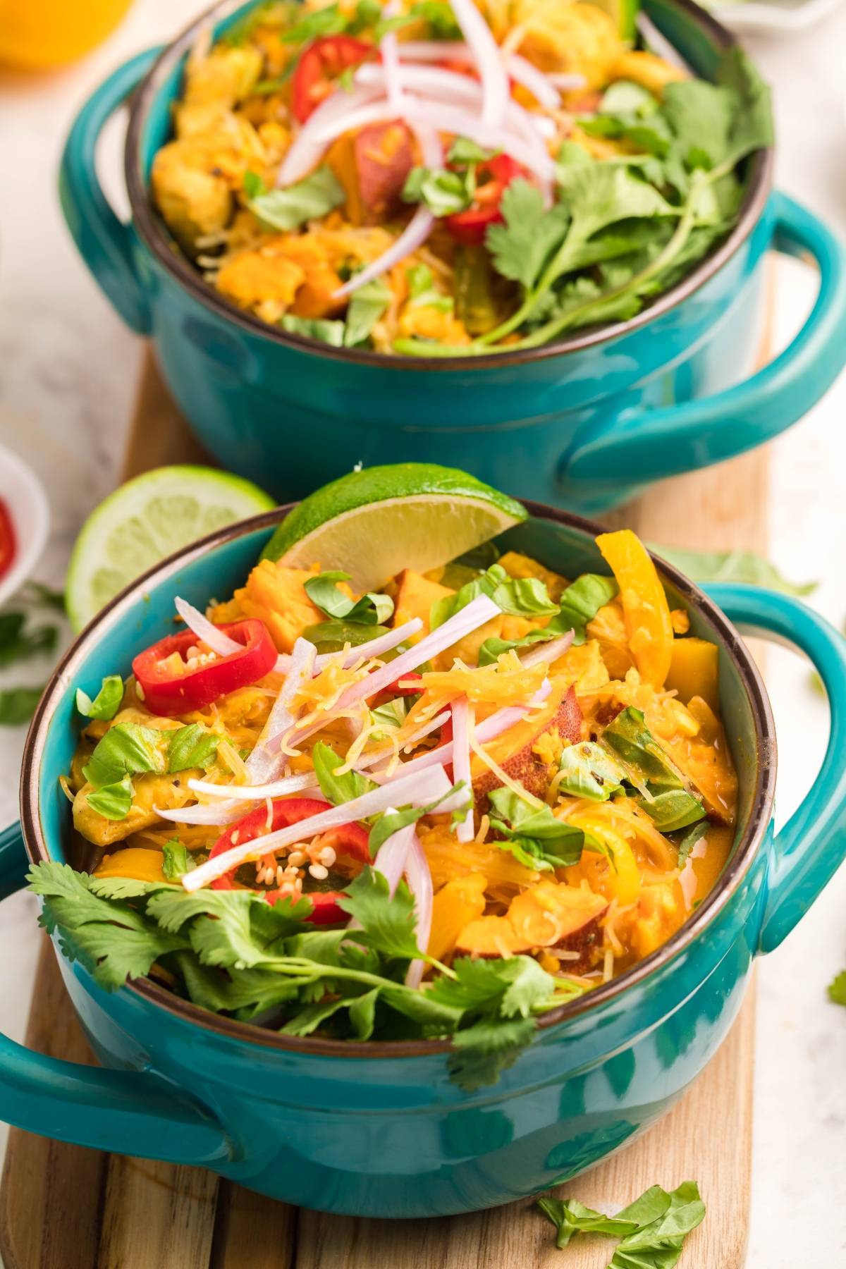 Two bowls of vibrant curry inspired by a hearty Thai recipe with chicken, topped with cilantro, lime wedges, red onions, and sliced chili peppers.