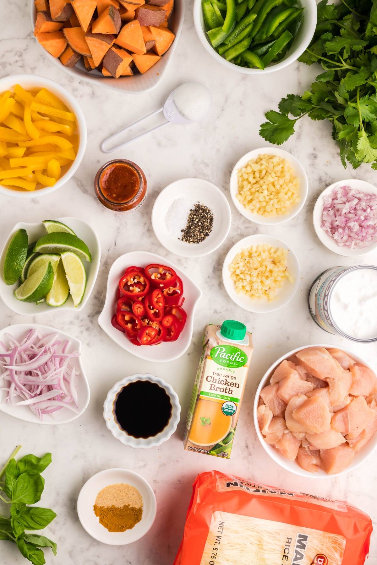 Various chopped vegetables, chicken, seasonings, and broth arranged in bowls on a white marble surface for a hearty Thai recipe with chicken.