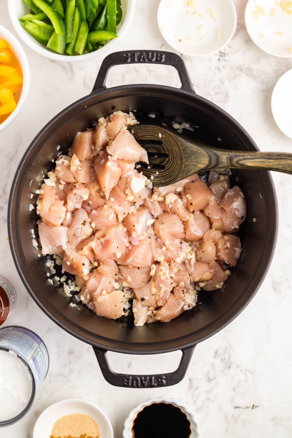 Diced raw chicken and chopped onions cooking in a black pot, surrounded by bowls of ingredients for a Thai chicken soup recipe with sweet potatoes, peppers, and red curry.