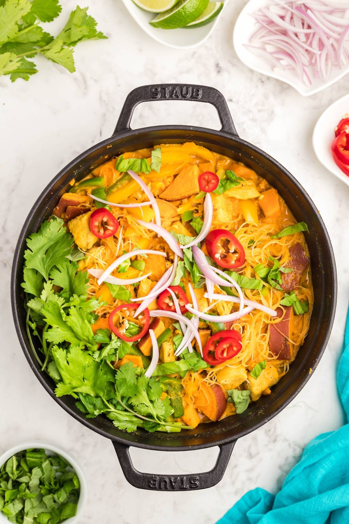 A black pot of Thai Chicken and Sweet Potato Soup with cilantro, red onion, and red chili slices sits on a white surface—a hearty Thai recipe with chicken and vibrant flavors.