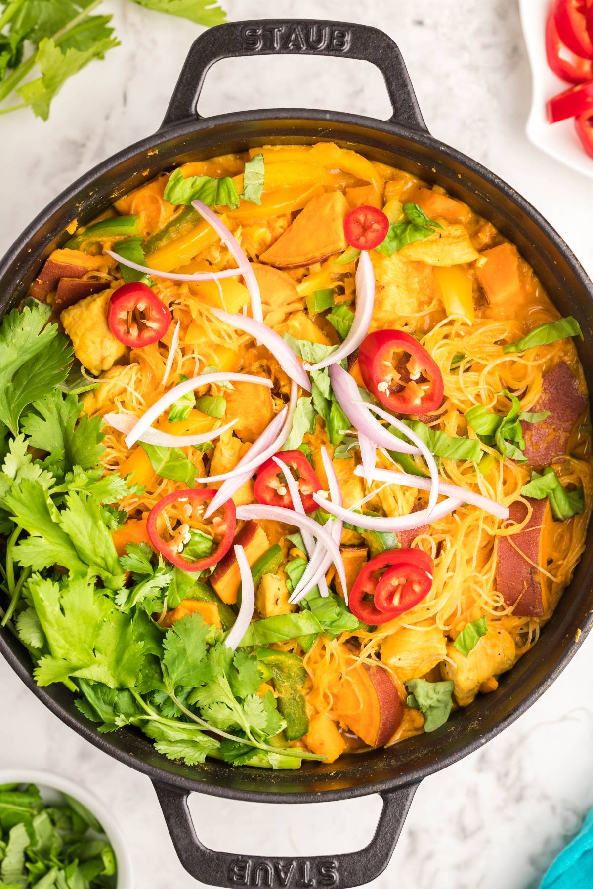 A pot of vibrant soup with red curry, noodles, sweet potatoes, cilantro, red chili, and sliced red onion on top.