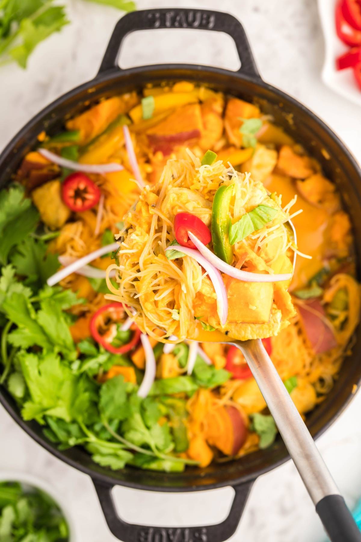 A ladle lifting curry noodles with tofu, sweet potatoes, peppers, and vegetables from a pot—a vibrant soup with sweet potatoes and peppers, garnished to perfection.
