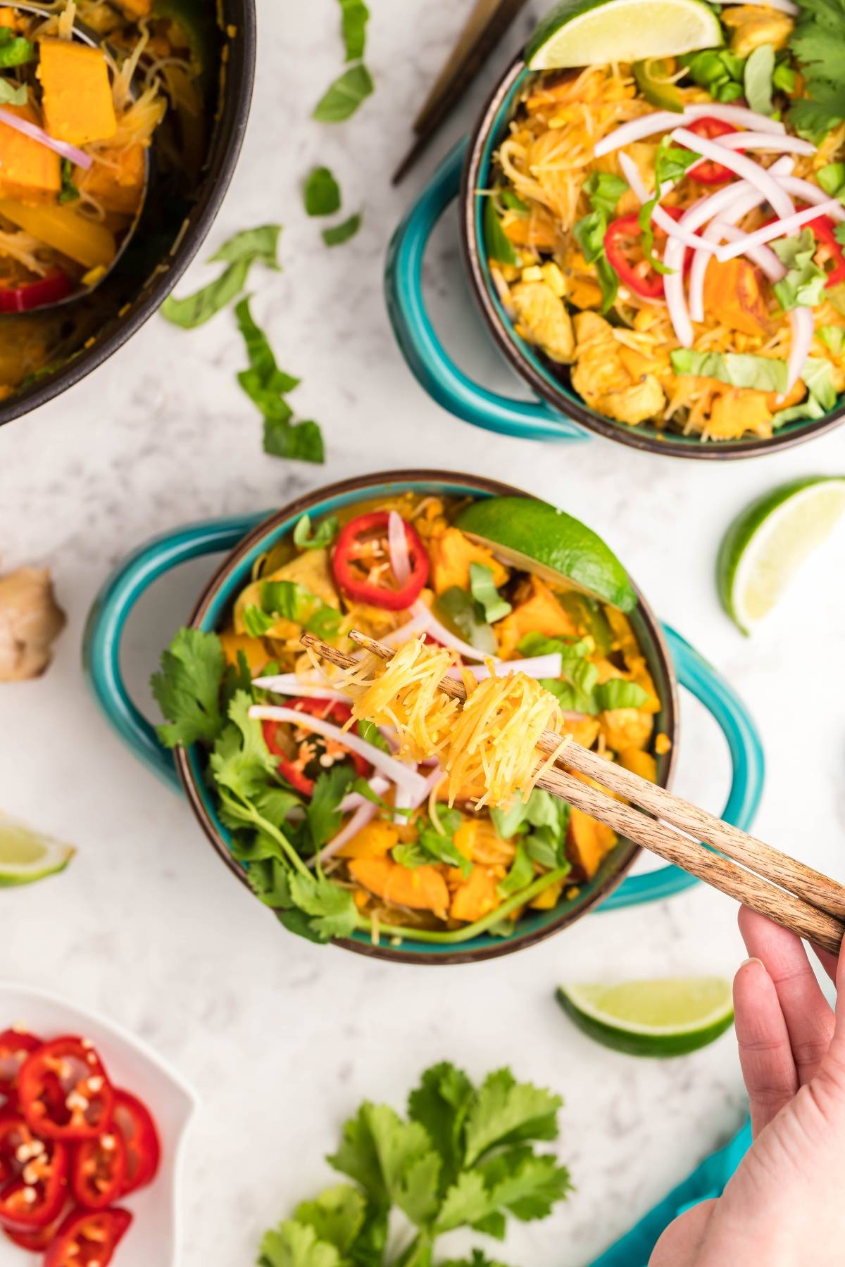 Overhead view of a bowl of Thai Chicken and Sweet Potato Soup with chicken, herbs, lime, and sliced vegetables, held with chopsticks.