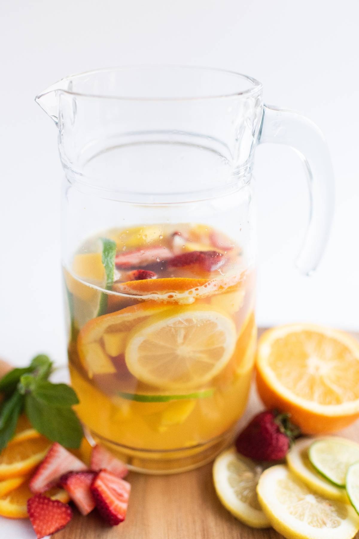 A glass pitcher of citrus and strawberry-infused water, surrounded by fresh fruit slices on a wooden surface—perfect as a refreshing summer drink.
