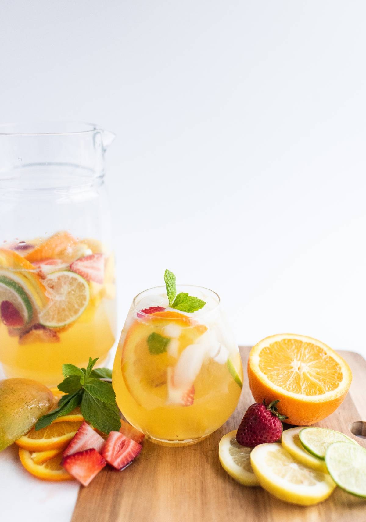 A glass and pitcher of white sangria with summer fruit, featuring citrus, berries, and mint, served with fresh slices on a wooden board.
