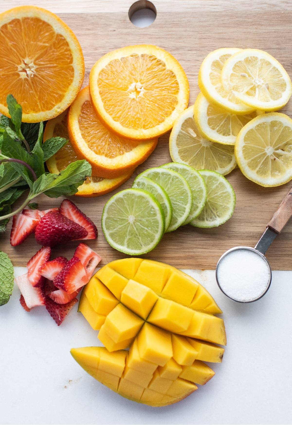 Sliced orange, lemon, lime, strawberries, cubed mango, mint, and a spoon of sugar on a cutting board—perfect ingredients for a refreshing white sangria with summer fruit.