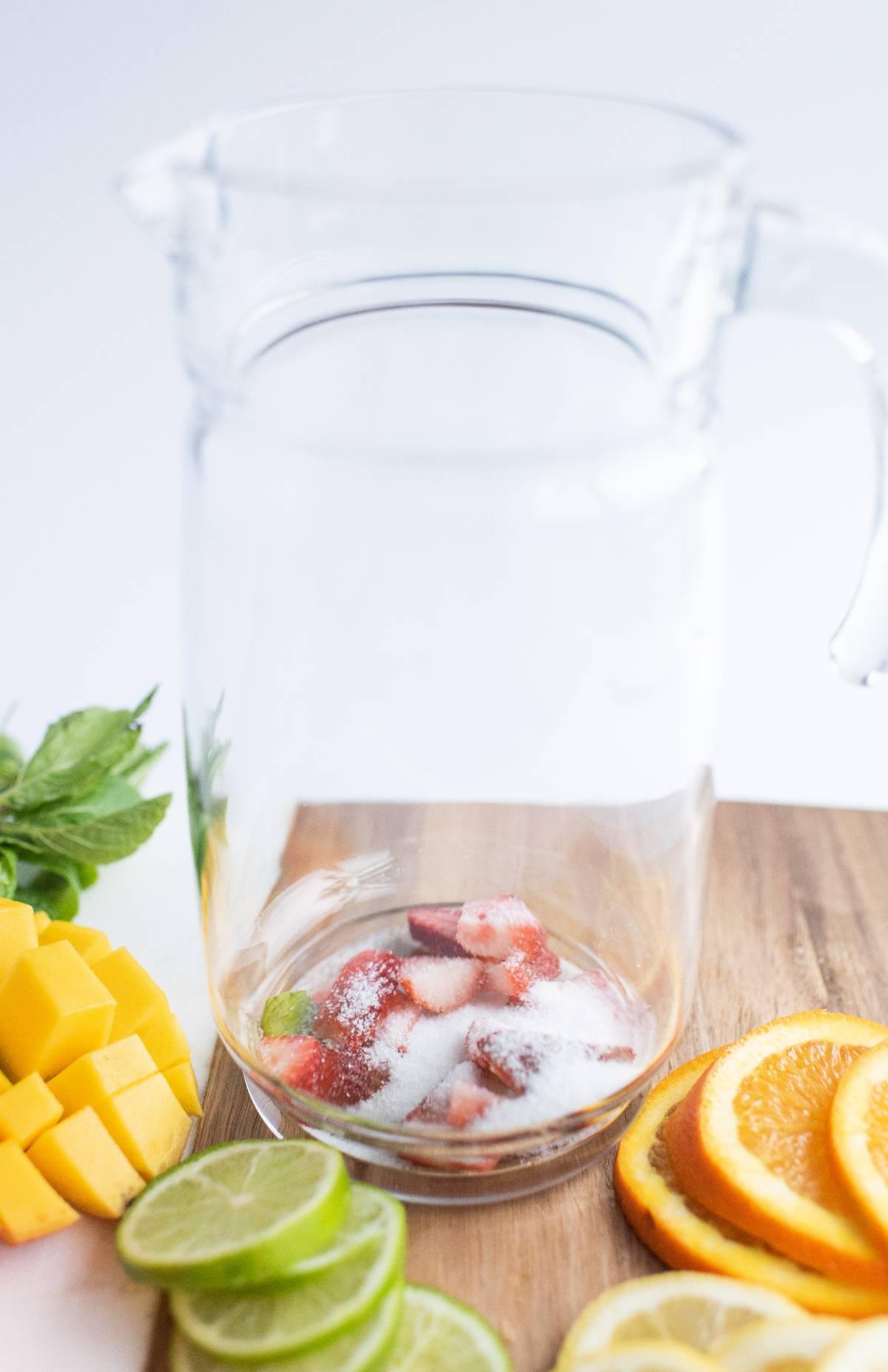 A glass pitcher with sliced strawberries and sugar, surrounded by mango, lime, lemon, orange, and mint—perfect for a refreshing summer drink or a vibrant white sangria-inspired summer fruit cocktail recipe.