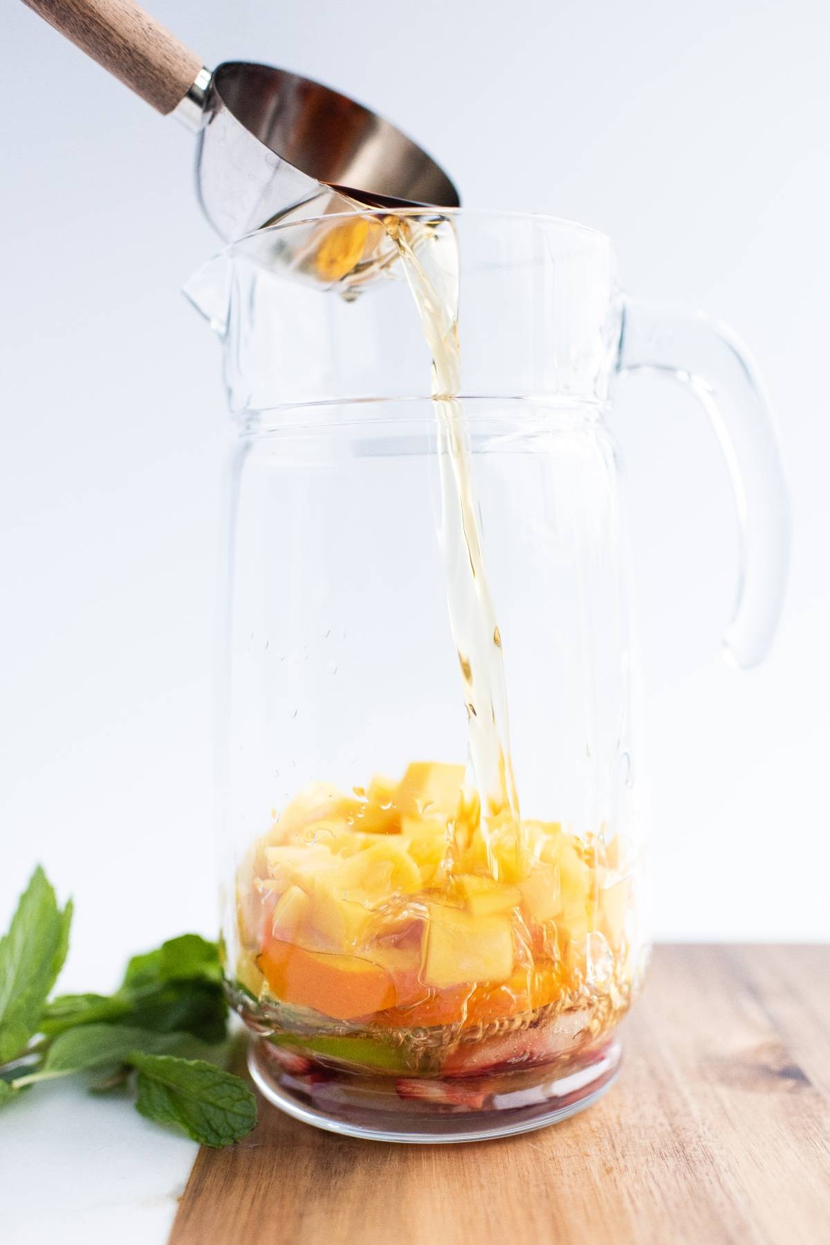 Iced tea being poured into a pitcher with fruit slices and mint on a wooden surface—perfect for a refreshing summer drink or as a super easy sangria recipe.
