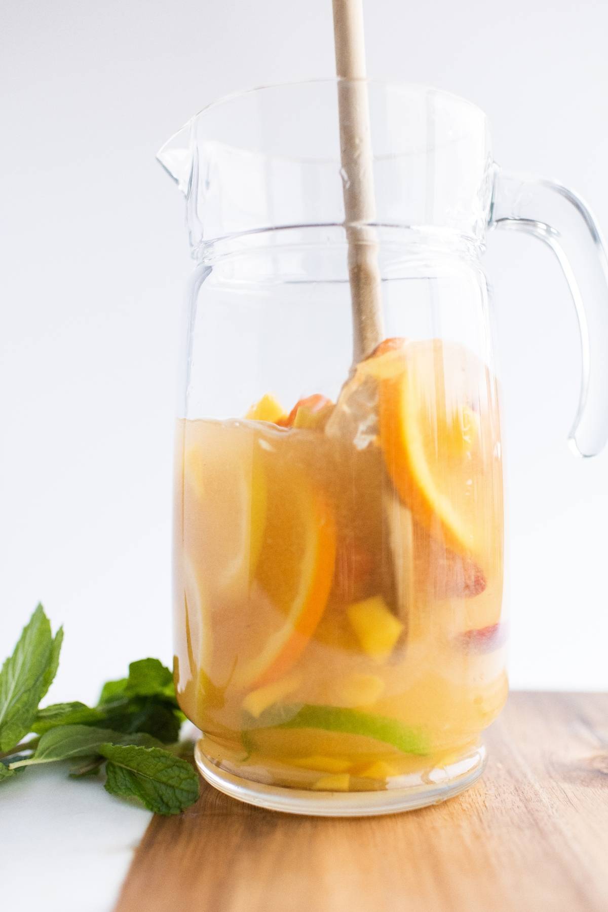 A glass pitcher of refreshing summer drink with orange slices and mint sits on a wooden surface, perfect for serving as a vibrant white sangria with summer fruit.